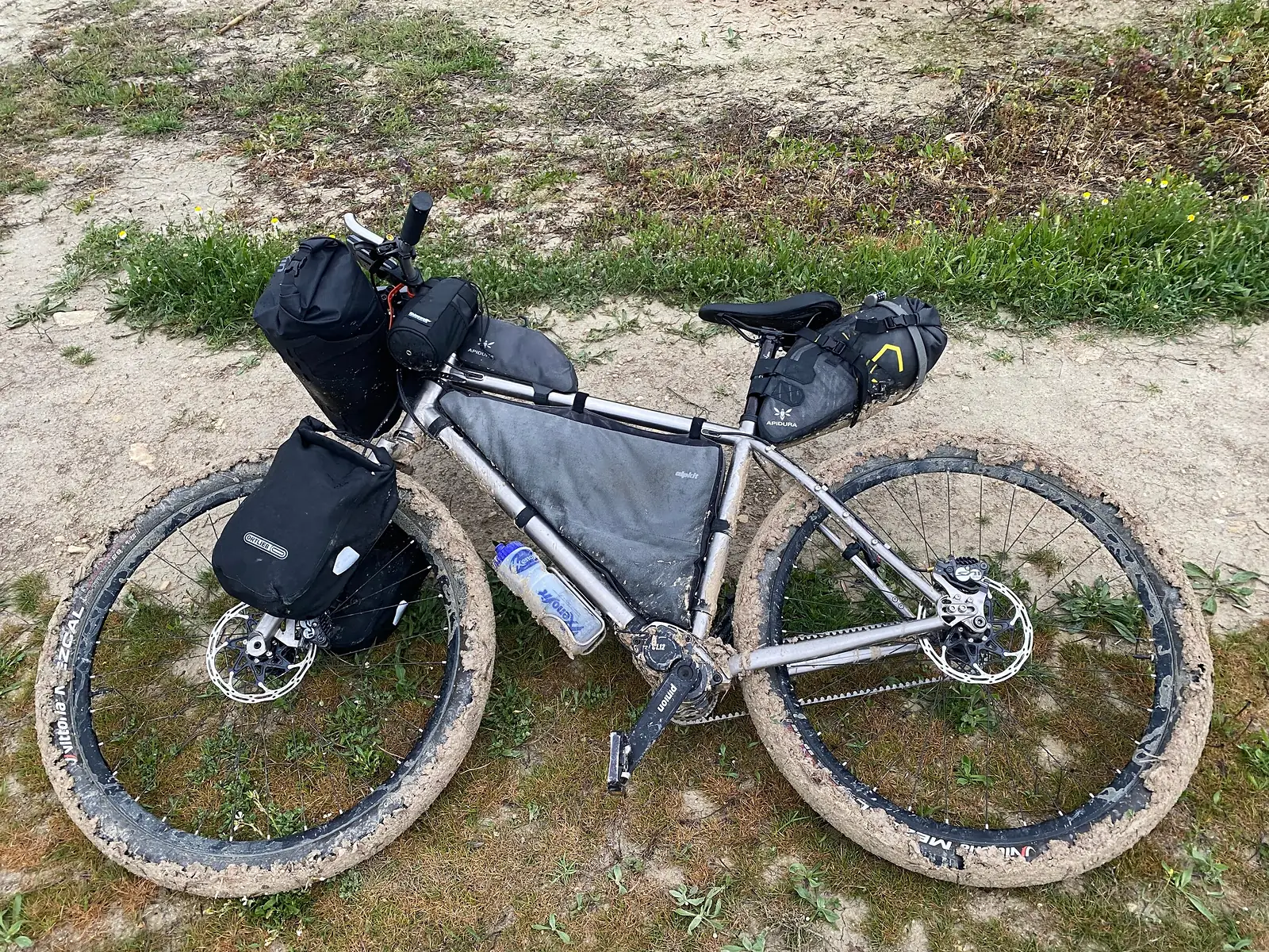  A loaded titanium adventure bike lying on its side on sandy grass, its tyres and rims packed solid with pale clay mud.