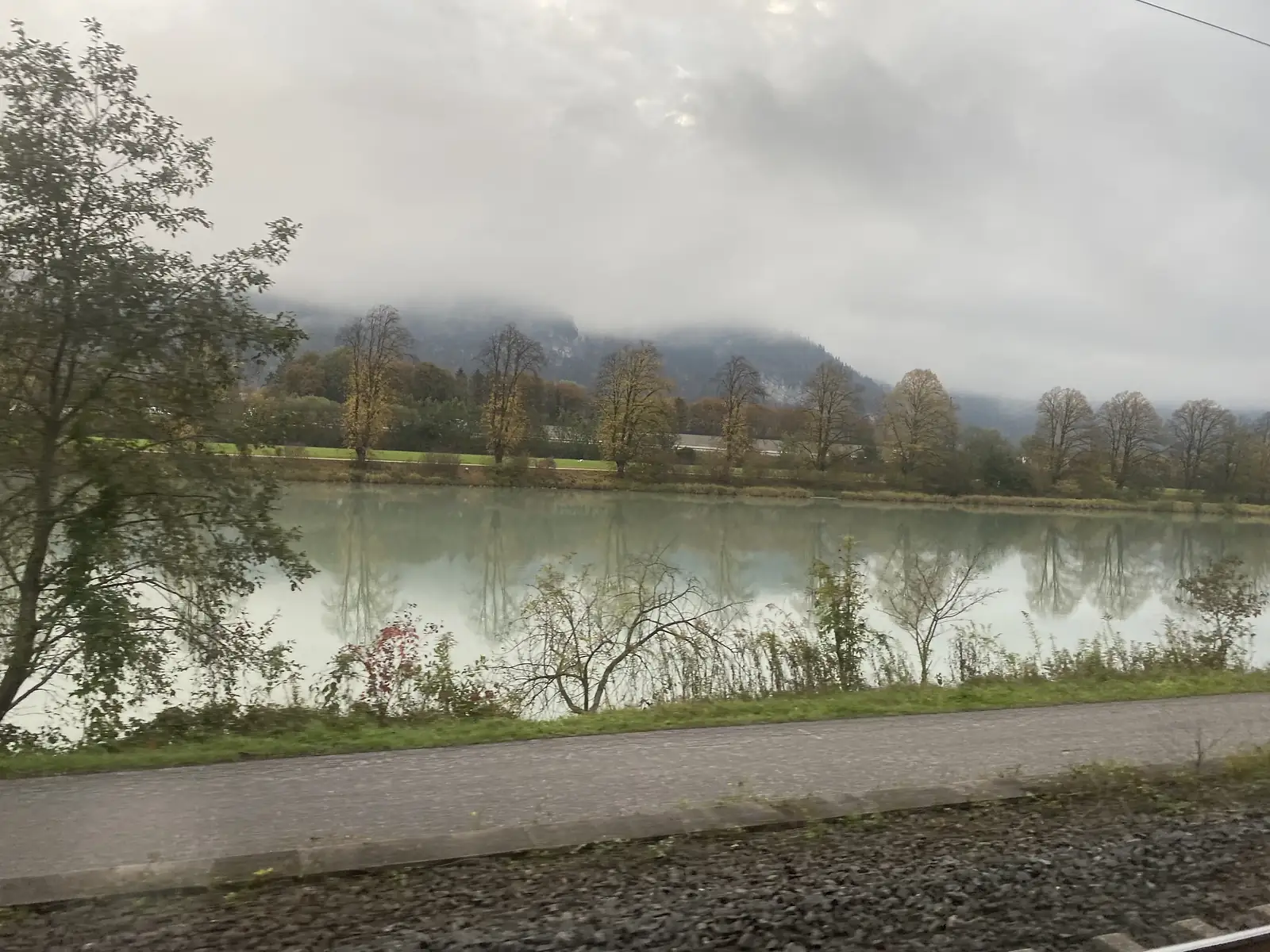 A calm river reflects a line of autumn trees, seen from a train window with low cloud clinging to the mountainside beyond.