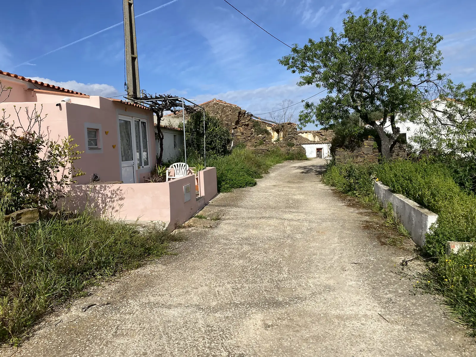 Concrete lane climbing through a hamlet past a pink-rendered house with a white chair outside, roofless stone ruins further along and an almond tree beside the road.