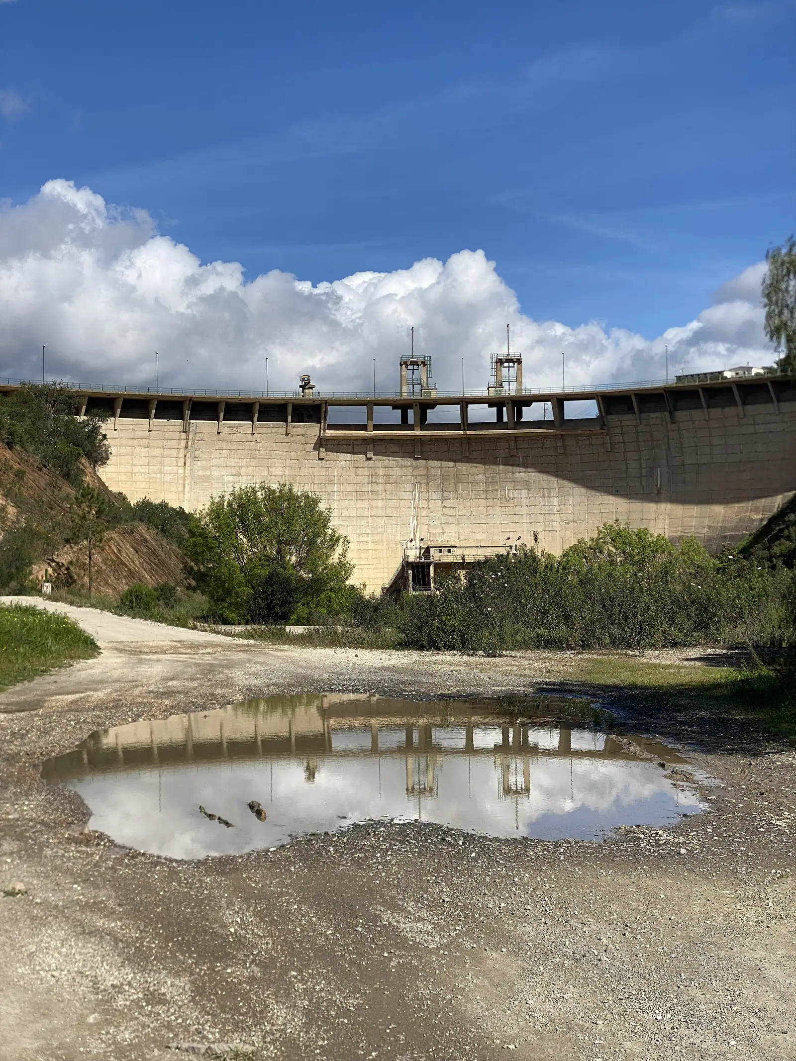 Curved concrete dam wall with winch towers along its crest, seen from the dry ground below and reflected whole in a large rain puddle in the foreground.