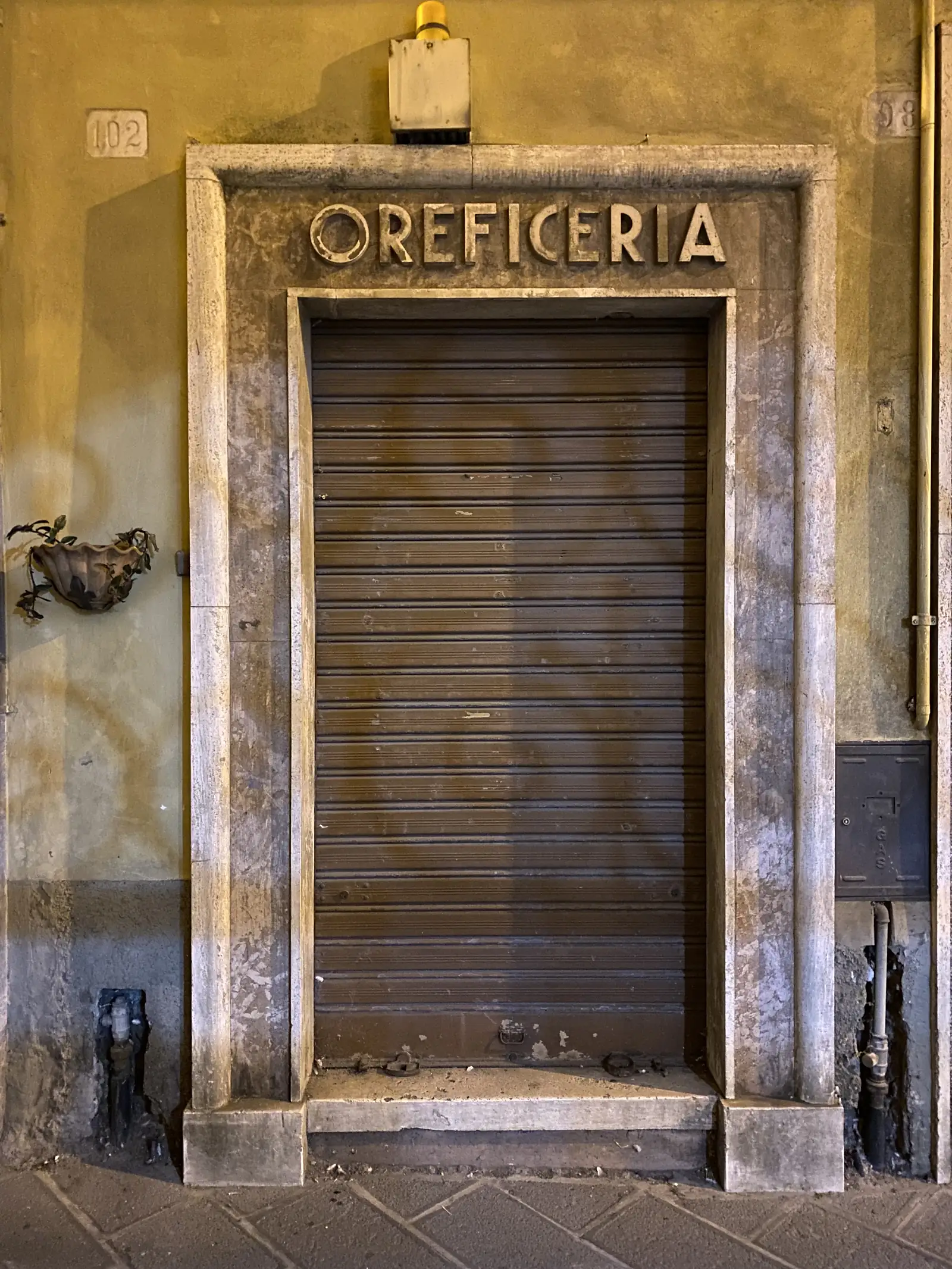 A closed metal shutter over a shopfront set in a stone surround carved with the word "OREFICERIA" (jeweller's), a small potted plant beneath.
