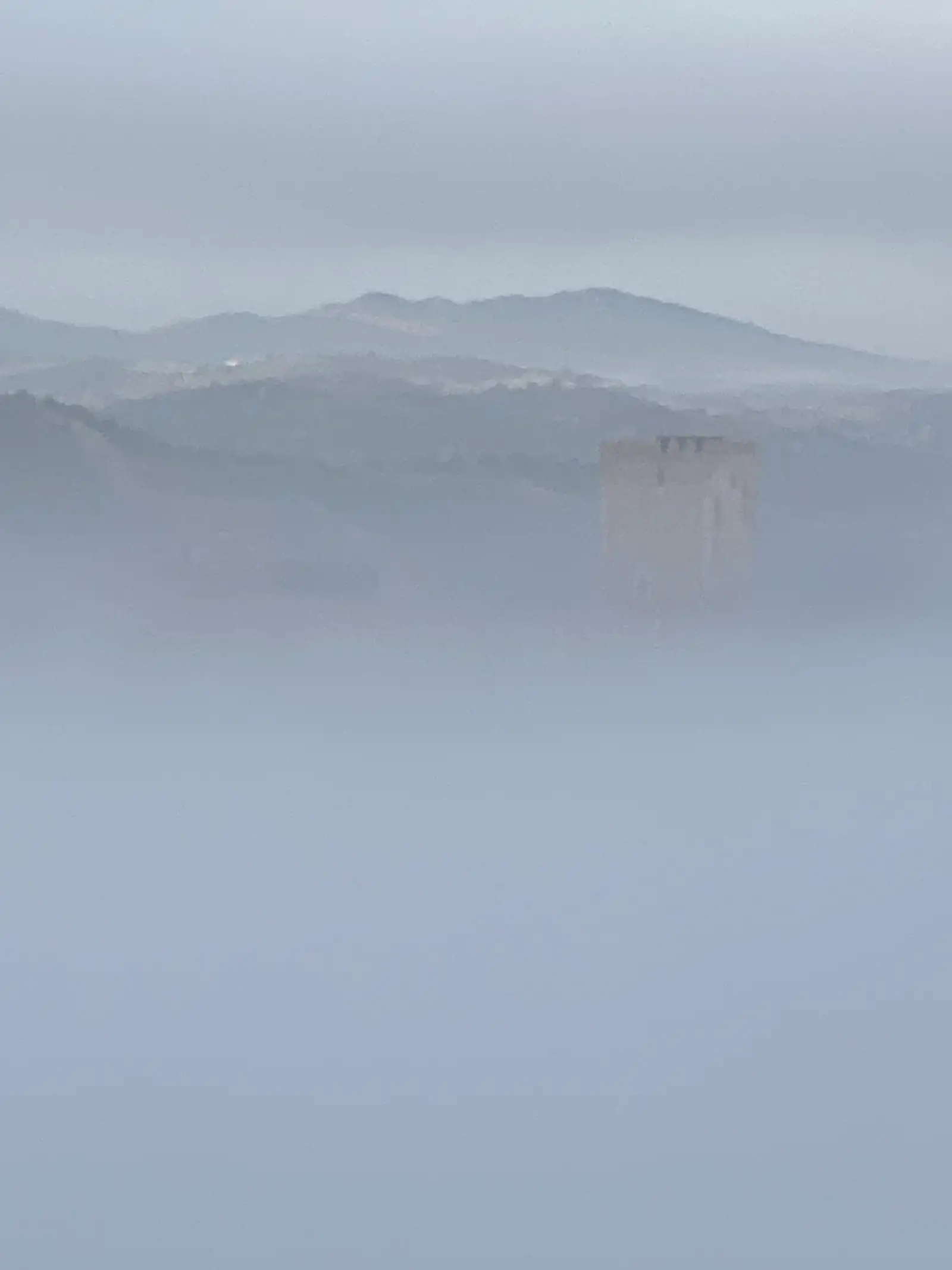 A crenellated castle keep rising out of a valley filled to the brim with fog, with wooded ridges and distant hills showing faintly beyond.