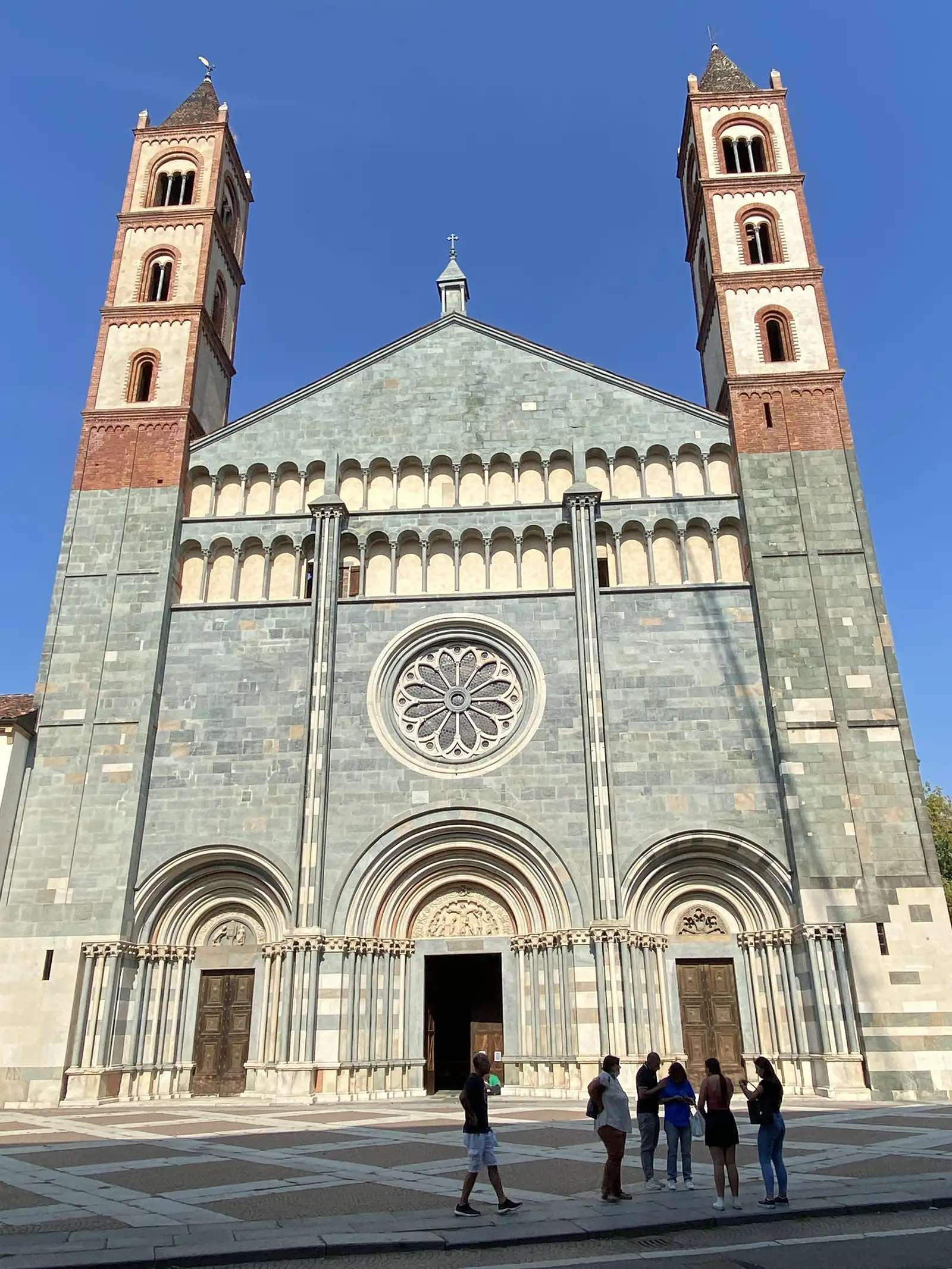 The basilica's bell towers seen across a lawned garden, with a bronze monument inscribed 'Umberto' standing among trees in the foreground.