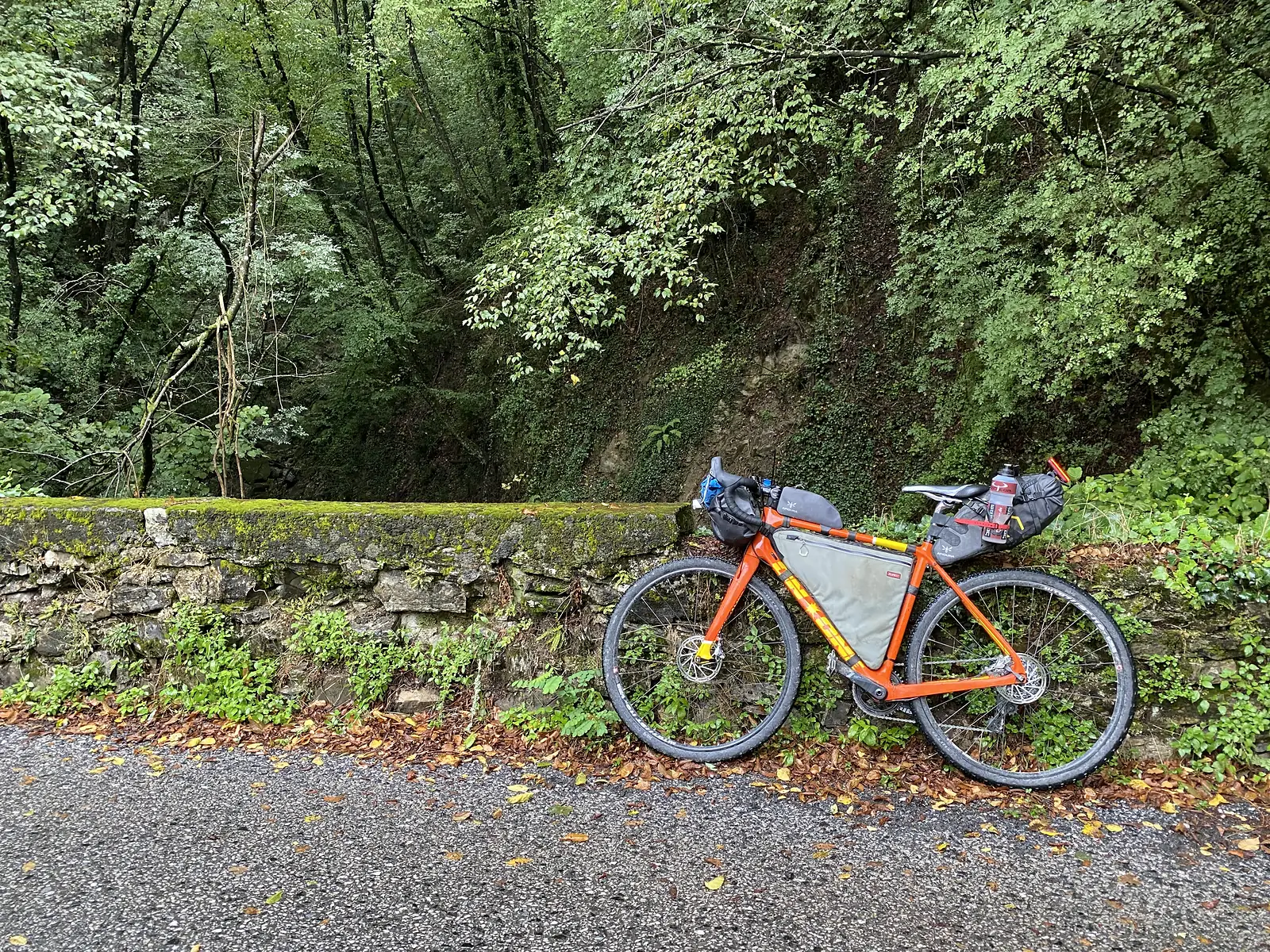 A loaded orange gravel bike with black frame bags and bottles leans against a moss-covered dry-stone wall beside a wooded lane.