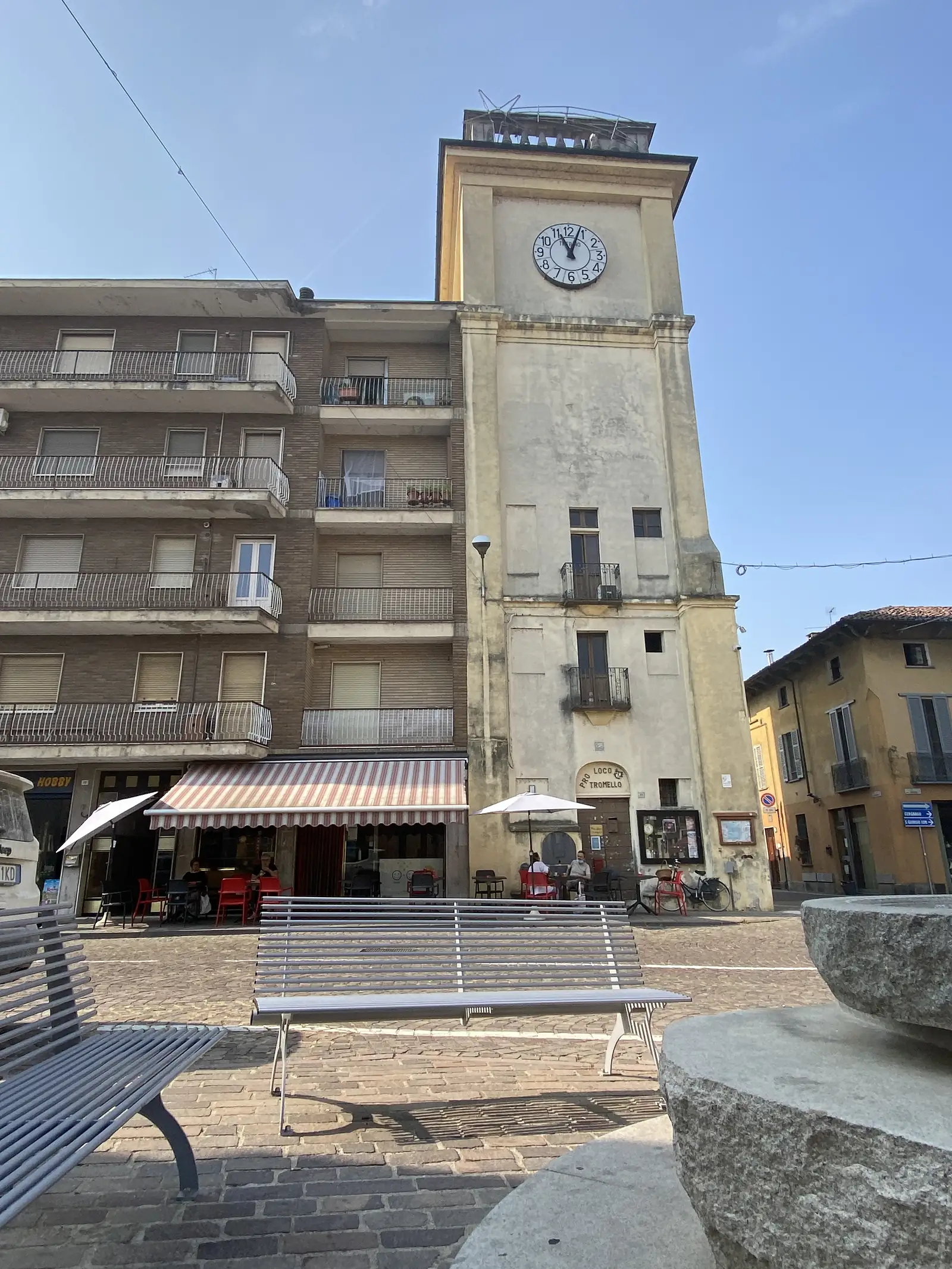 A stone clock tower rises above a small-town square, its base carrying a sign reading 'PRO LOCO TROMELLO' beside a café with red chairs under awnings.
