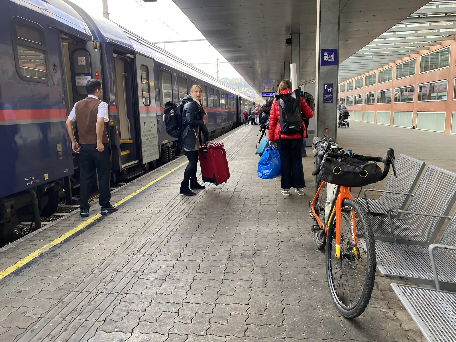 Passengers with suitcases and rucksacks wait beside a couchette carriage marked 'Hamburg-Altena', while a loaded orange gravel bike stands parked by a bench on the platform.