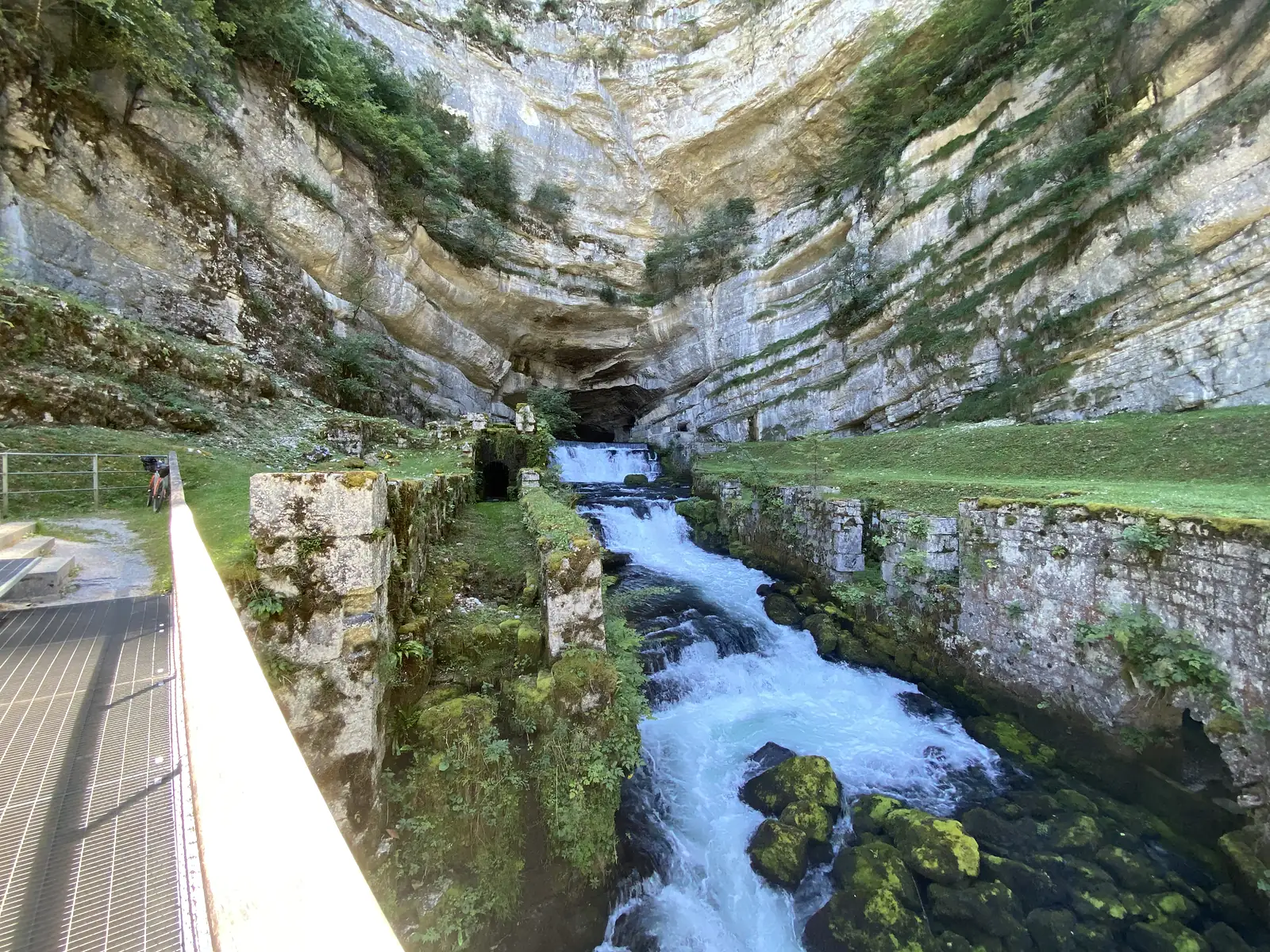 A spring gushes as a waterfall from a cave mouth at the head of a limestone amphitheatre, its water channelled between old stone walls.