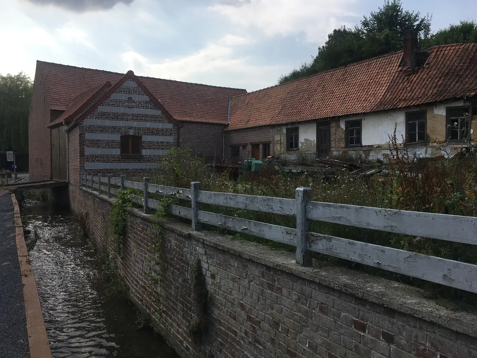 Weathered brick farm buildings with red-tiled roofs line a narrow stream behind a low brick wall and white post-and-rail fence.