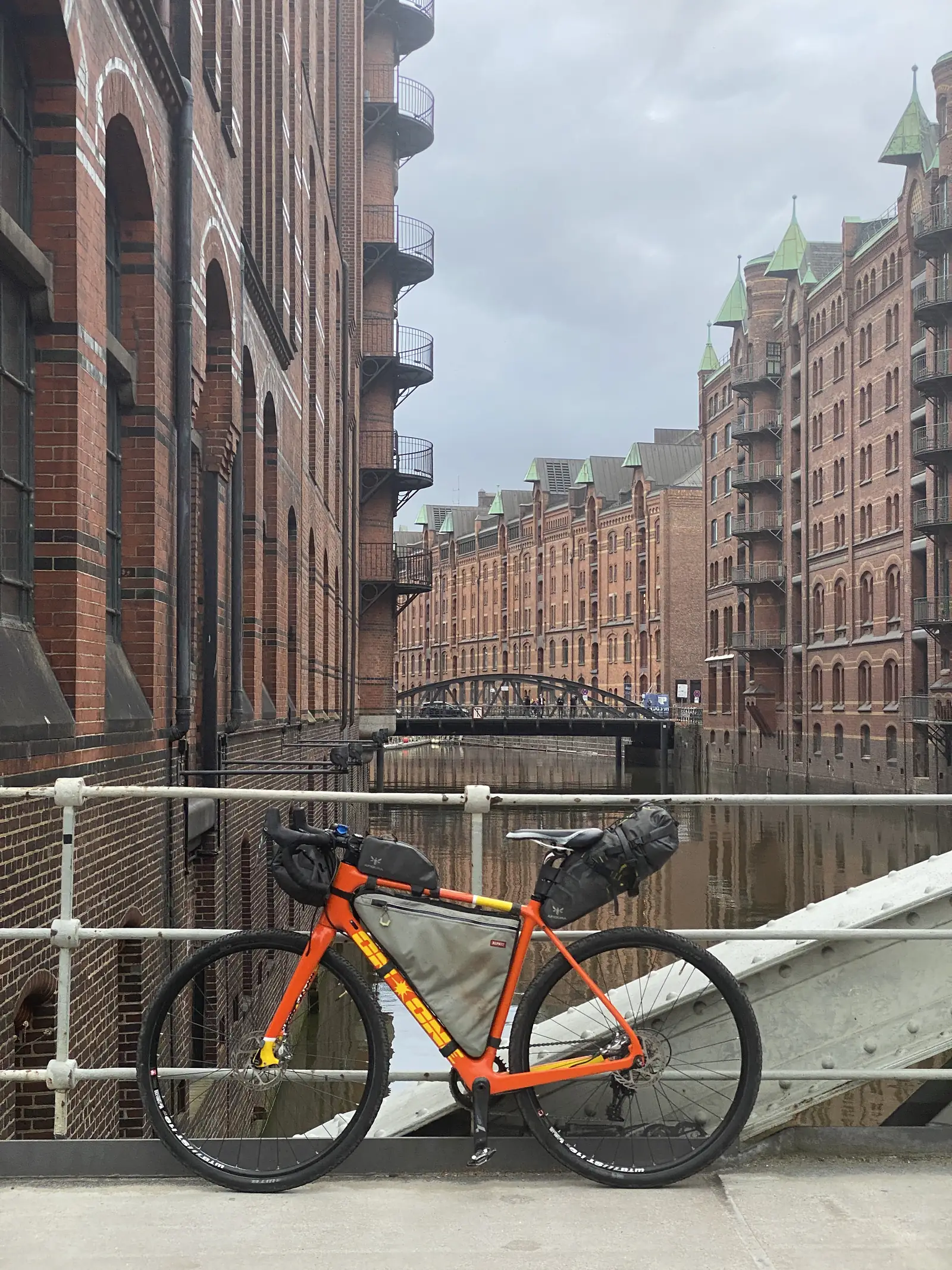 A loaded gravel bike leans against a railing on a bridge, with rows of red-brick warehouse buildings lining the canal behind it.