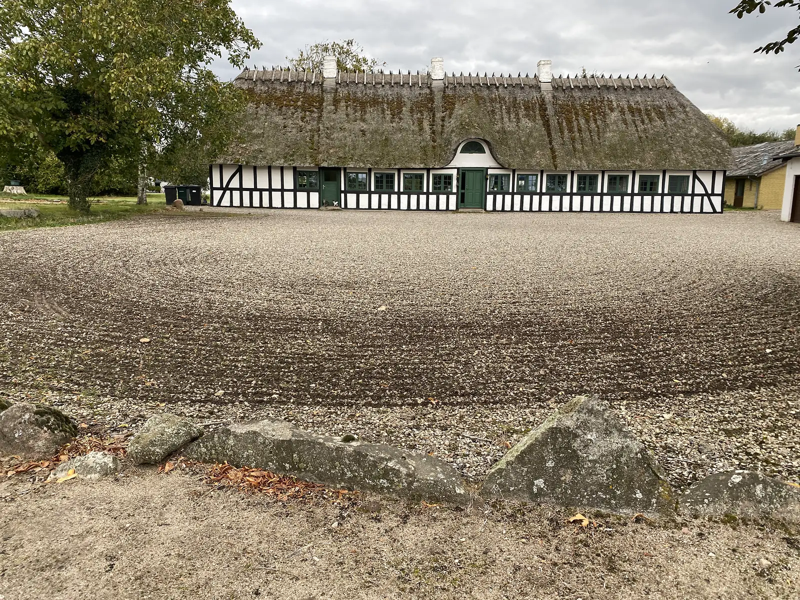 A long thatched half-timbered farmhouse with black timbers and green doors standing across a broad gravel yard.