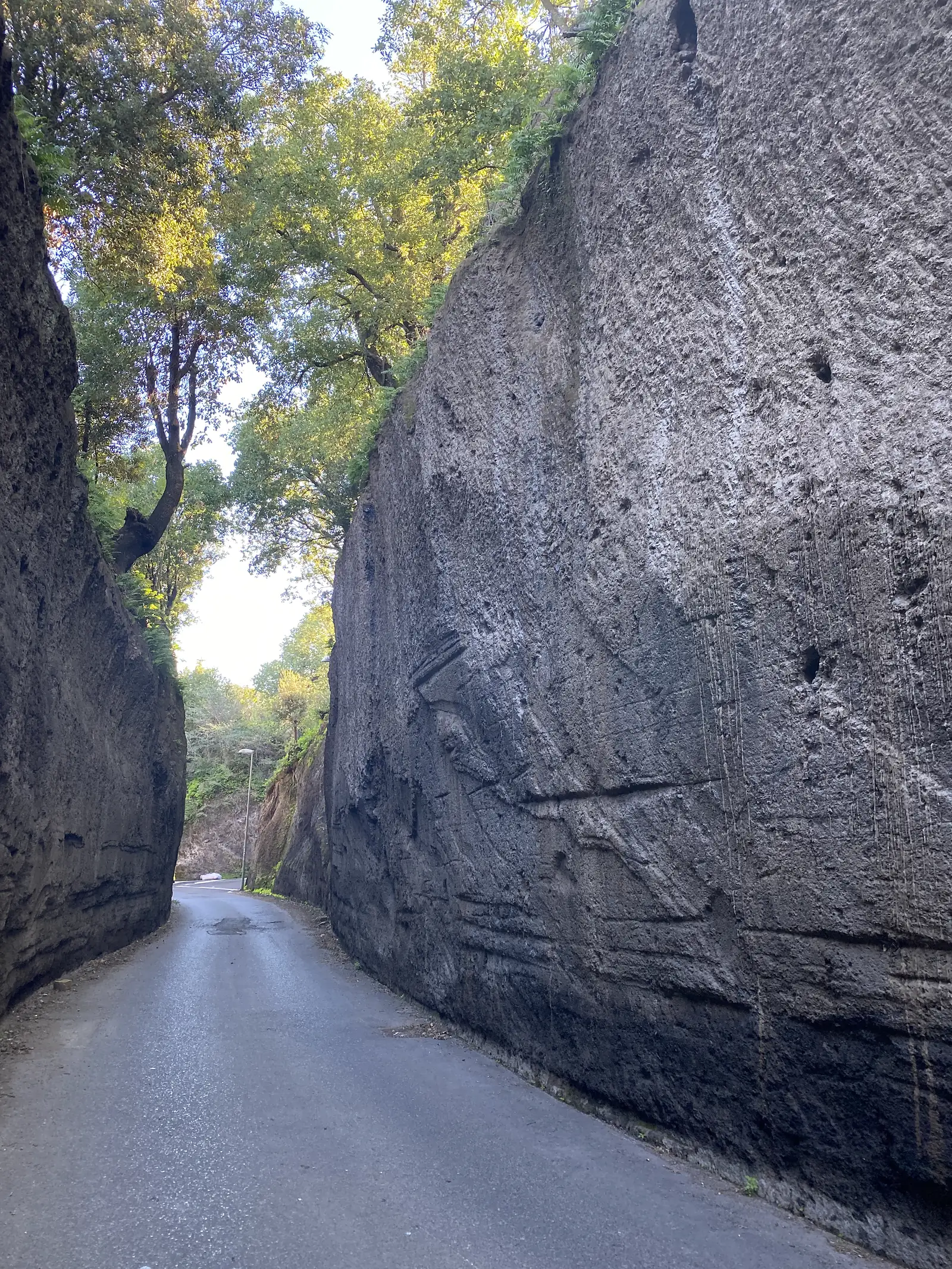 A narrow tarmac lane hemmed by tall tool-marked tufa rock walls, trees closing overhead further along.