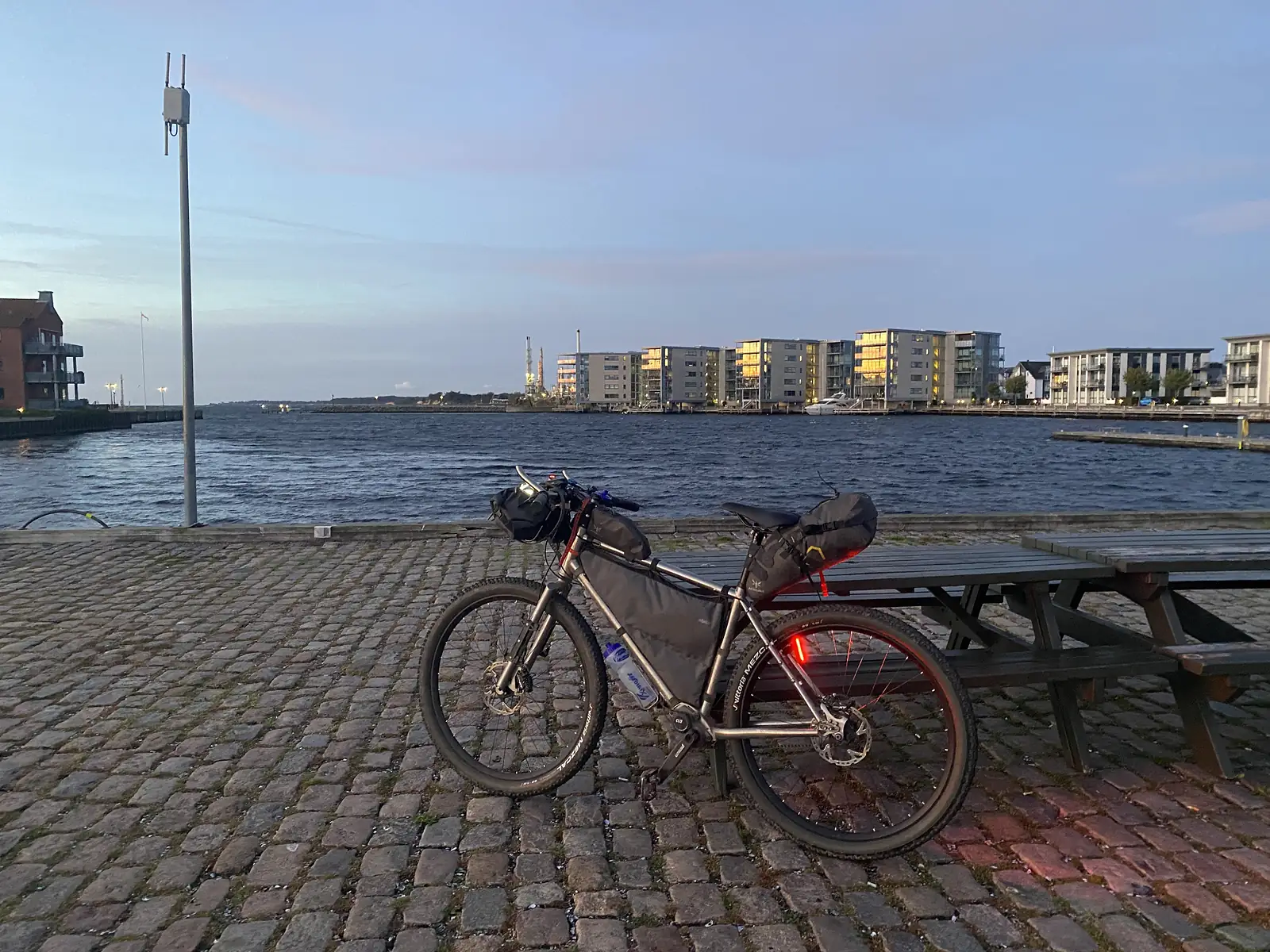 A loaded bikepacking bike standing on a cobbled quayside beside picnic tables at dusk, with modern waterfront apartment blocks across the water.