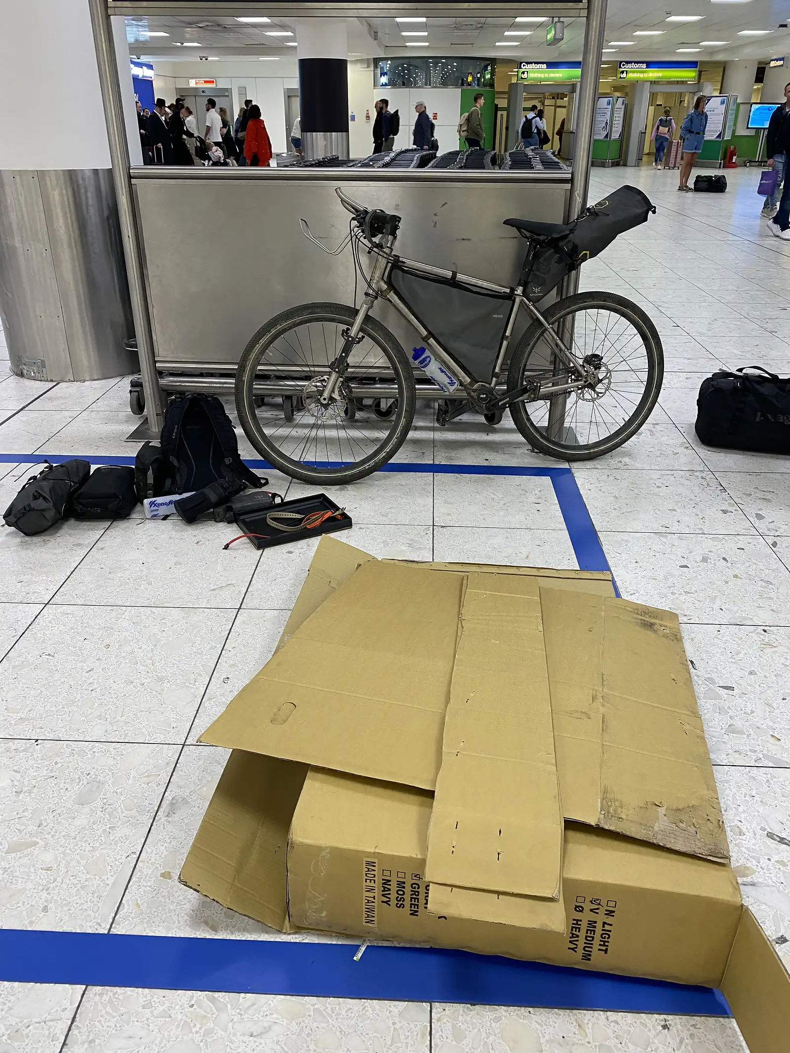 A rebuilt titanium bike propped against a baggage carousel in an airport arrivals hall, with flattened cardboard and tools on the floor.
