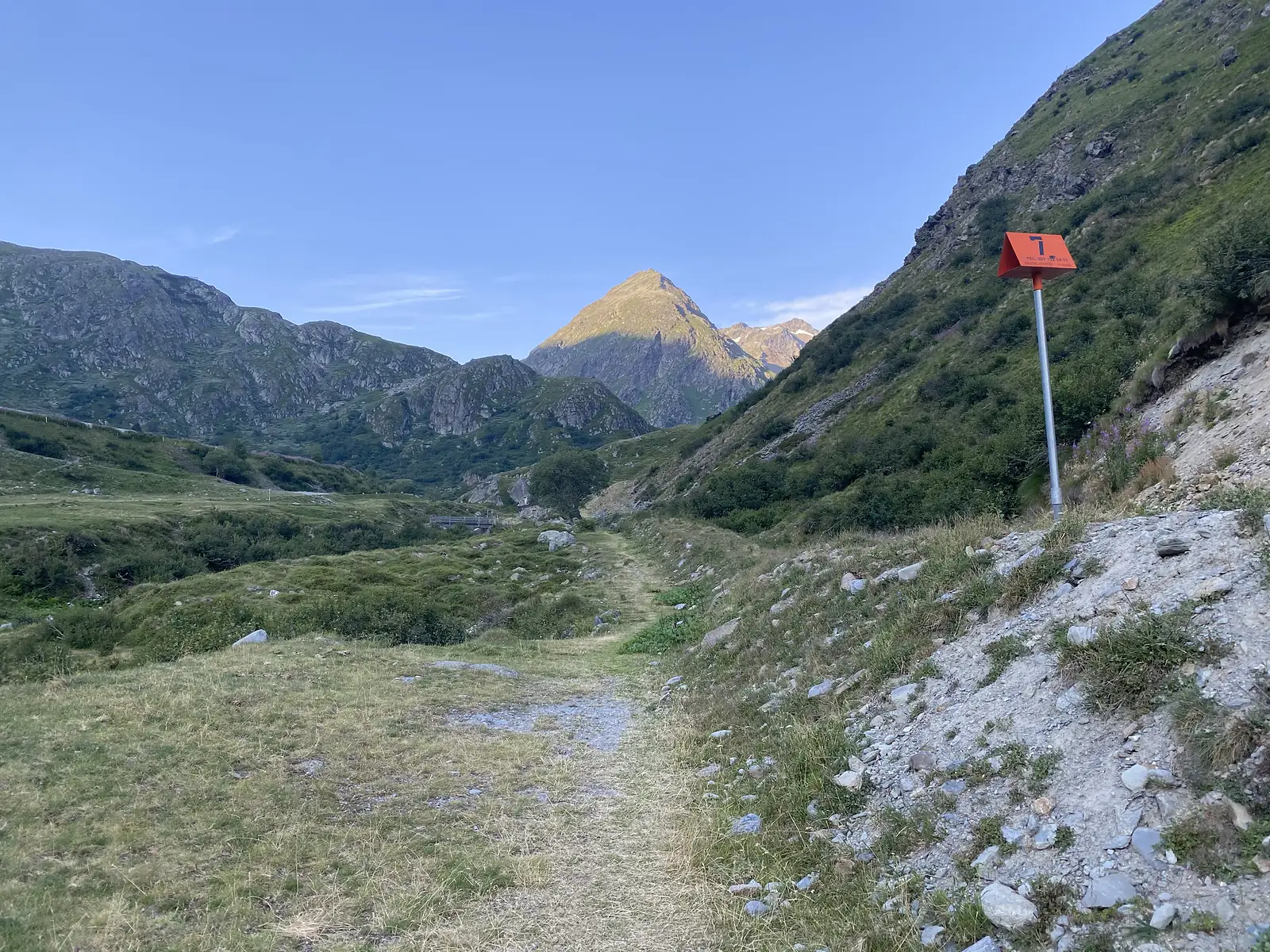 A stony path leads towards distant peaks past an orange numbered trail marker post reading '7'.