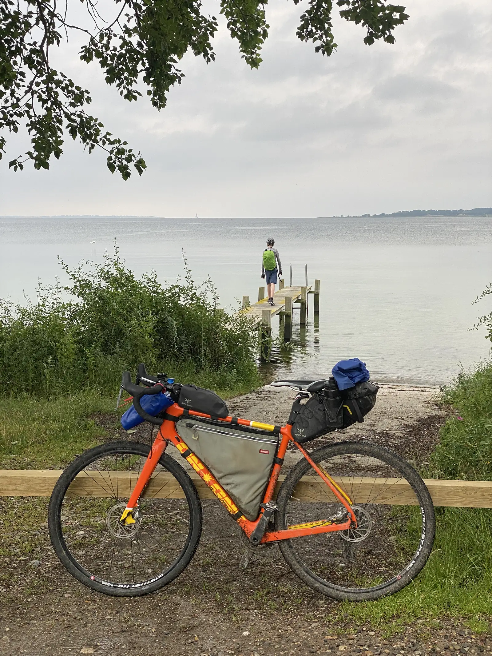 A loaded orange gravel bike propped against a rail above a shingle shore, with a young cyclist walking out along a timber jetty into a calm grey sea.