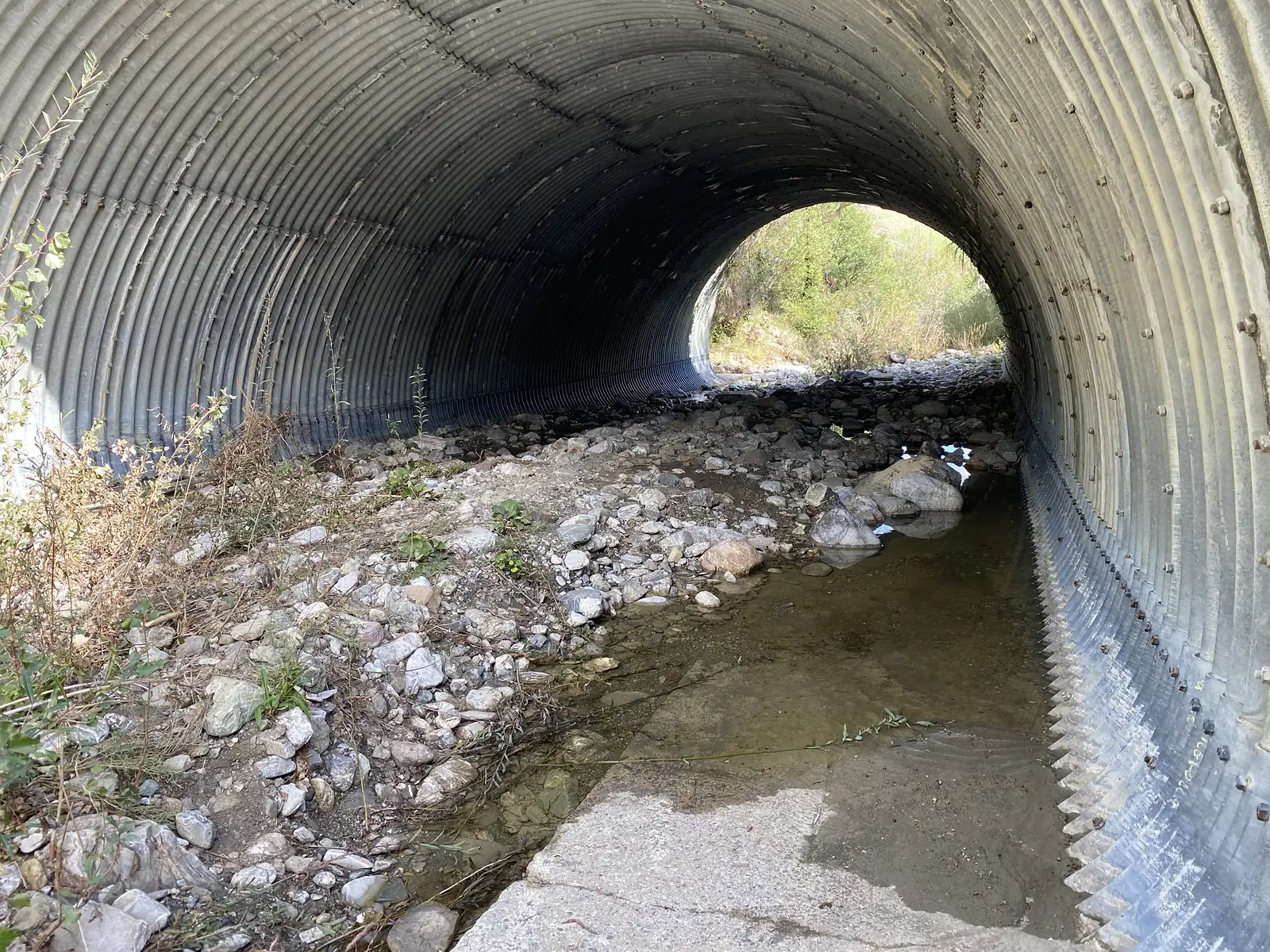 The inside of a large corrugated-metal culvert with a rocky, shallow stream running through it towards a bright wooded exit.
