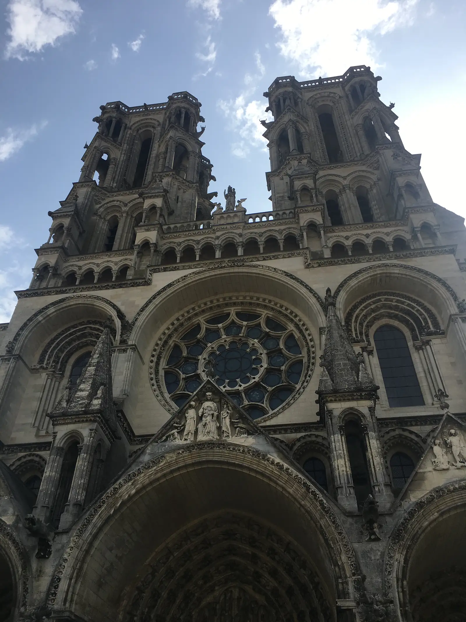 Two Gothic cathedral towers, decorated with tracery and gargoyles, stand against a partly cloudy sky above a rose window.