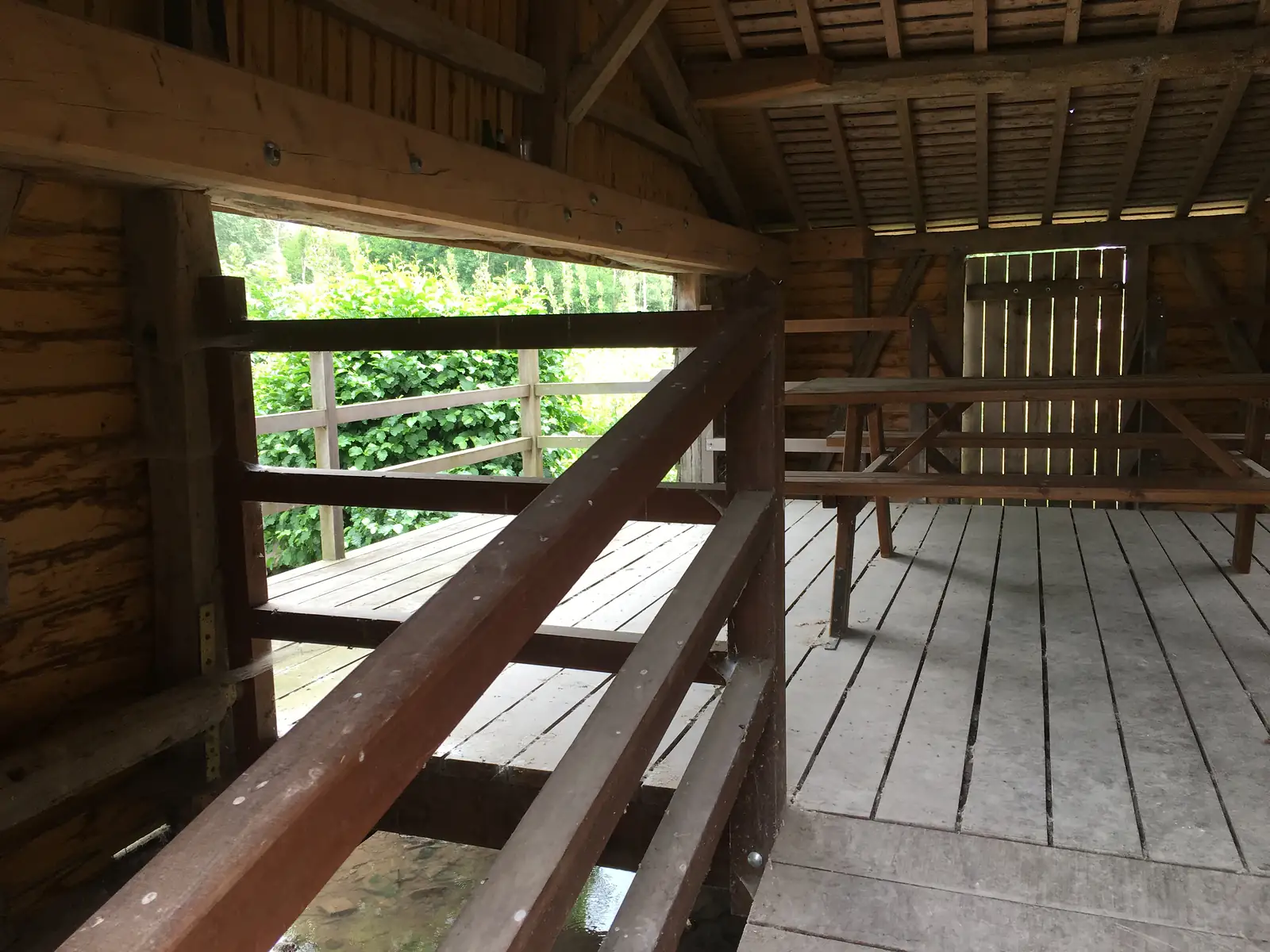 Inside the wooden shelter, a picnic table and benches sit on a decked floor, with a railed opening looking out over a hedge and stream below.