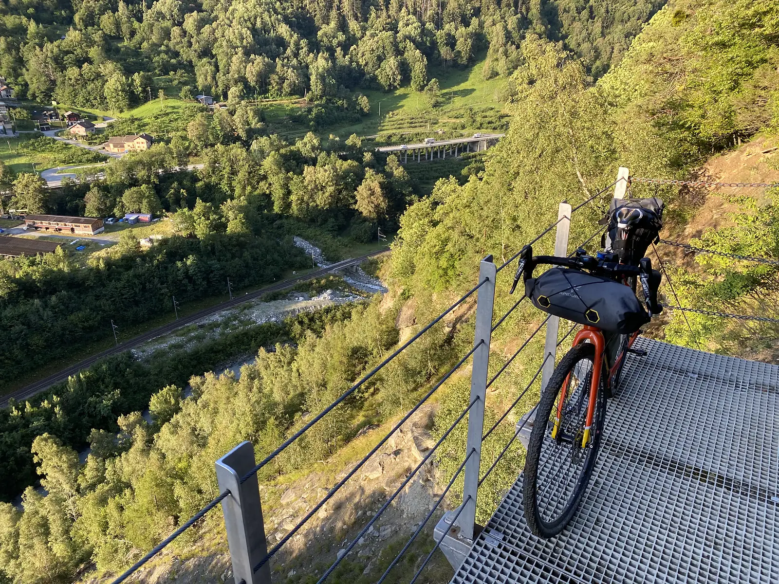 A red touring bike fitted with frame and saddle bags parked on a steel-grating viewing platform cantilevered over a wooded gorge, with a railway line, river and road viaduct far below.