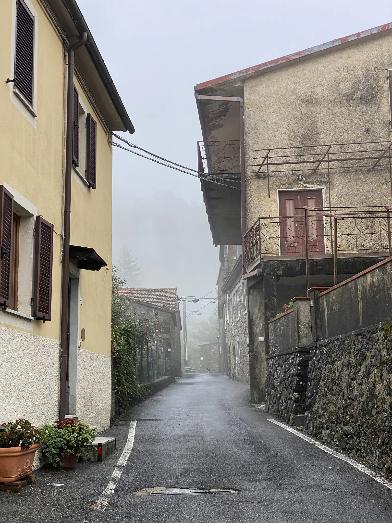 A narrow lane between rendered village houses with ornate iron balcony railings leads towards a misty archway further down the street.