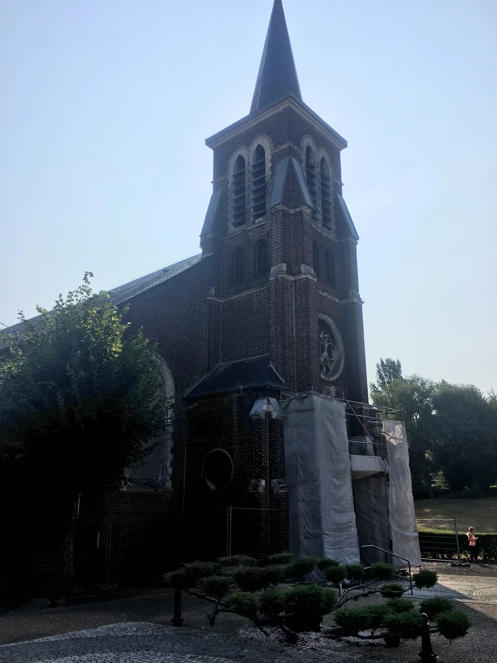 A tall brick church spire with pointed lancet windows and a rose window, part of the tower under scaffolding and sheeting during restoration work.