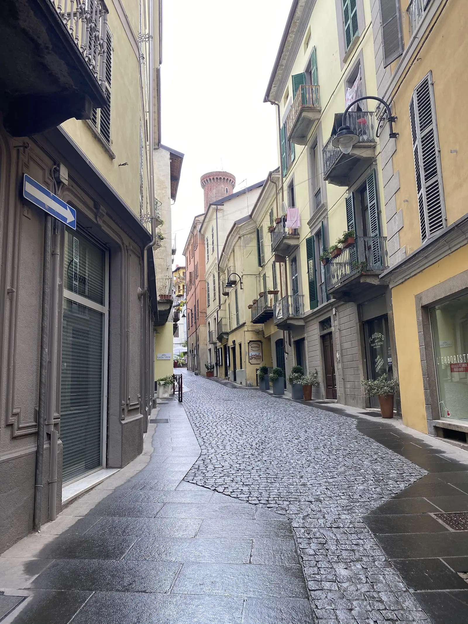 A cobbled street lined with yellow and terracotta townhouses leads towards a round brick tower rising above the rooftops of Ivrea's old town.