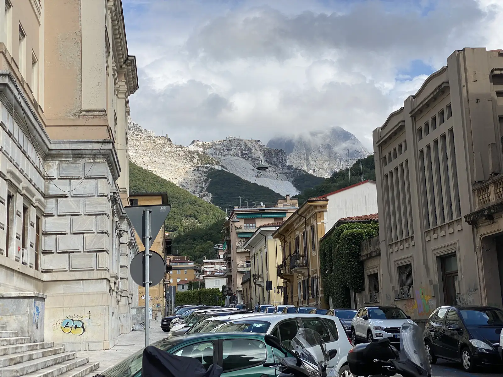 Marble quarry workings streak a distant mountain above a row of parked cars and grand old stone-faced buildings on a graffiti-marked street corner.