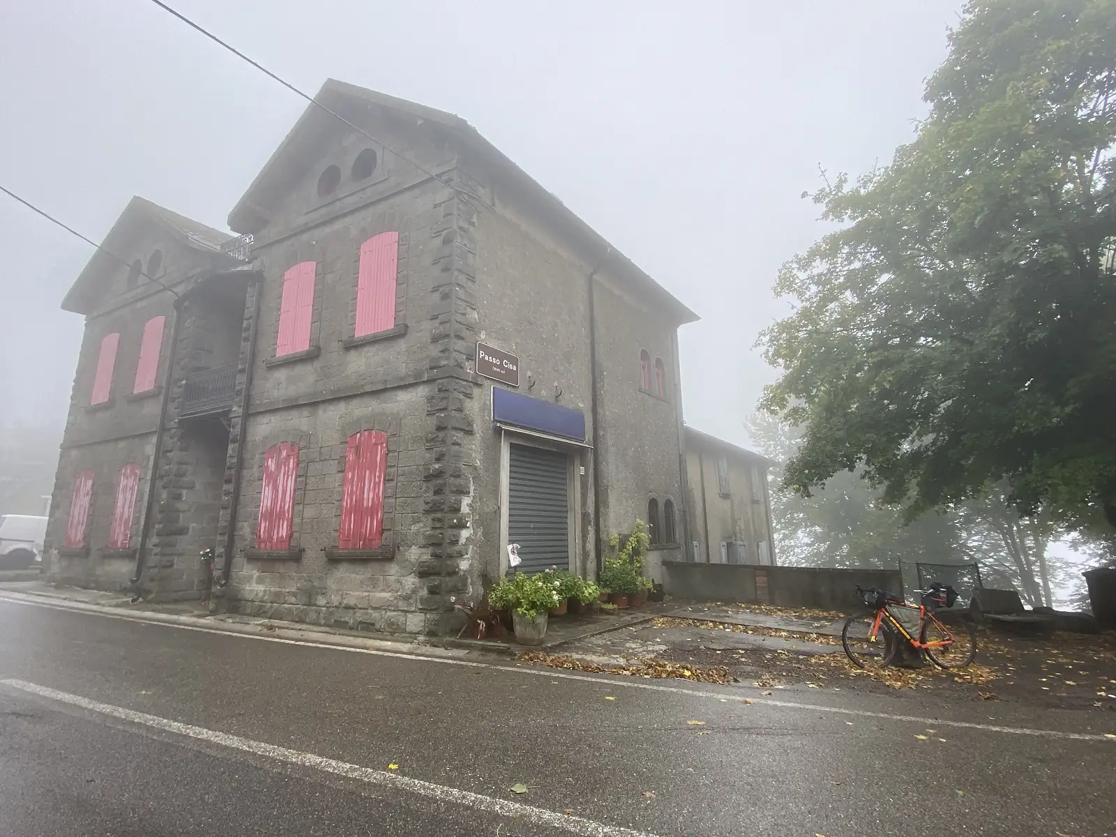 A stone building with faded pink shutters and a small sign reading Passo Cisa stands over a laden bike parked by the roadside in thick fog.