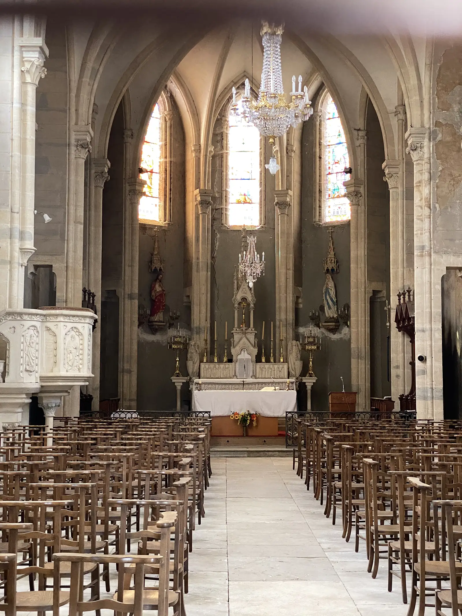 Rows of empty wooden chairs line the nave of a Gothic village church, leading past a stone pulpit to a candlelit altar beneath tall stained-glass windows and a crystal chandelier.
