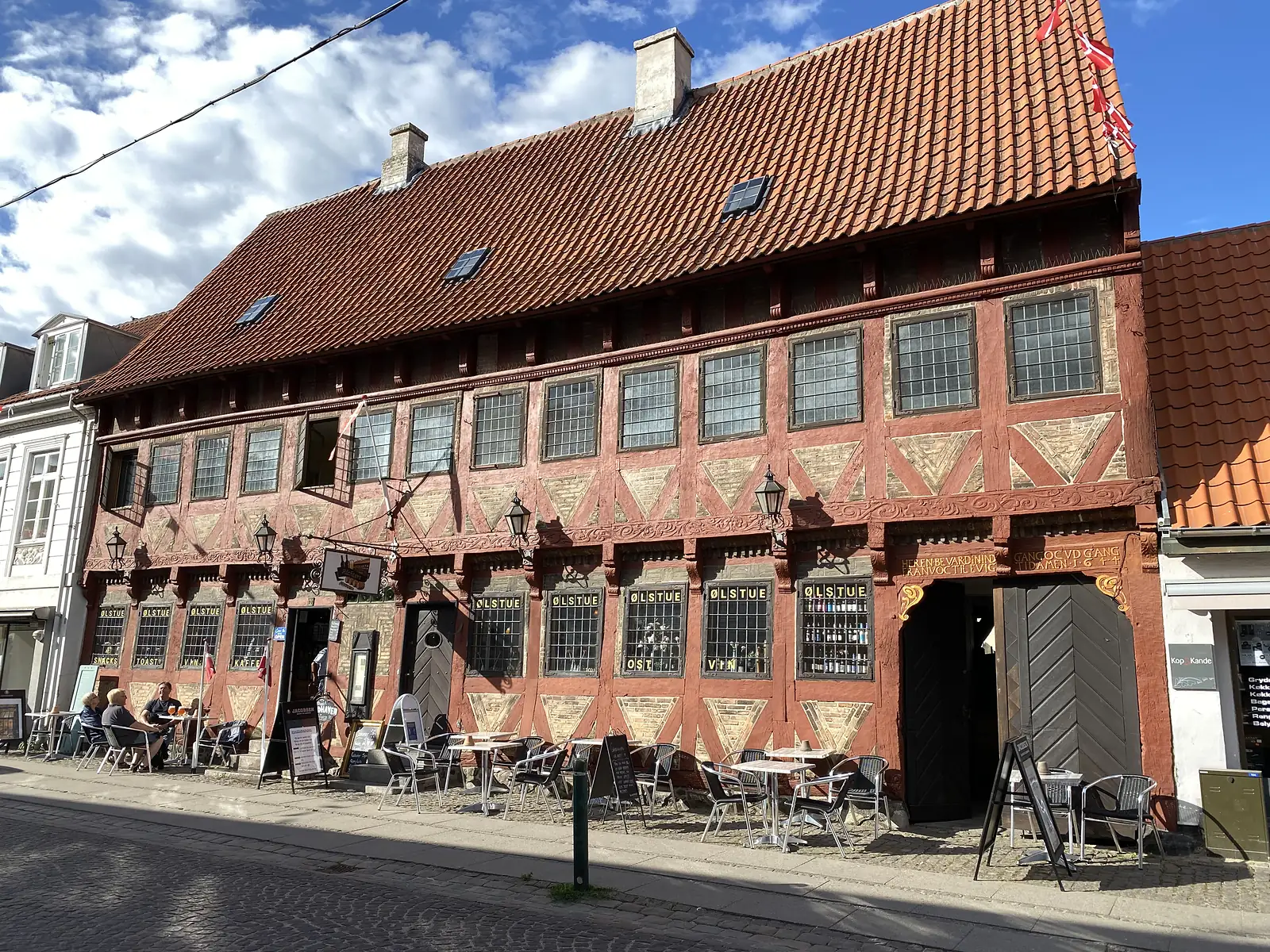 A long red half-timbered merchant's house with carved beams and leaded windows lettered "ØLSTUE", cafe tables on the pavement outside.