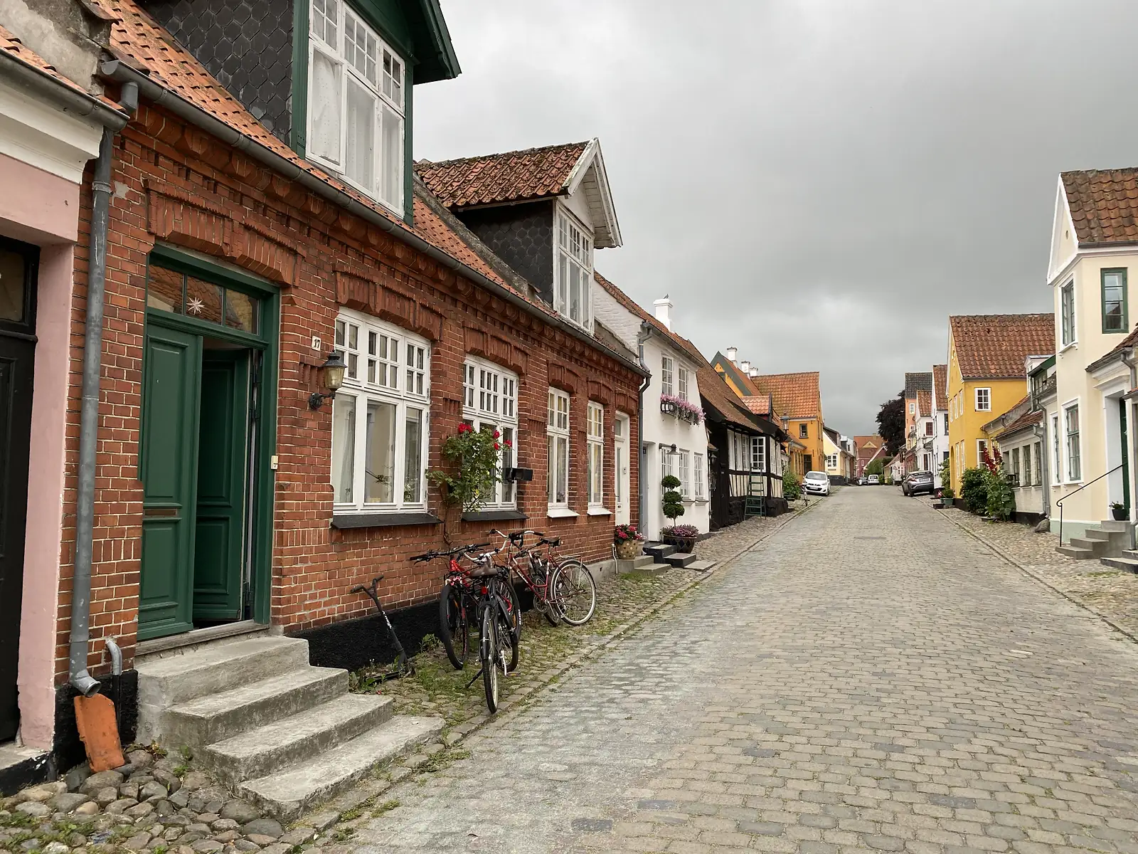 A cobbled street of red-brick and rendered houses with dormers and bicycles propped outside, curving away under a grey sky.