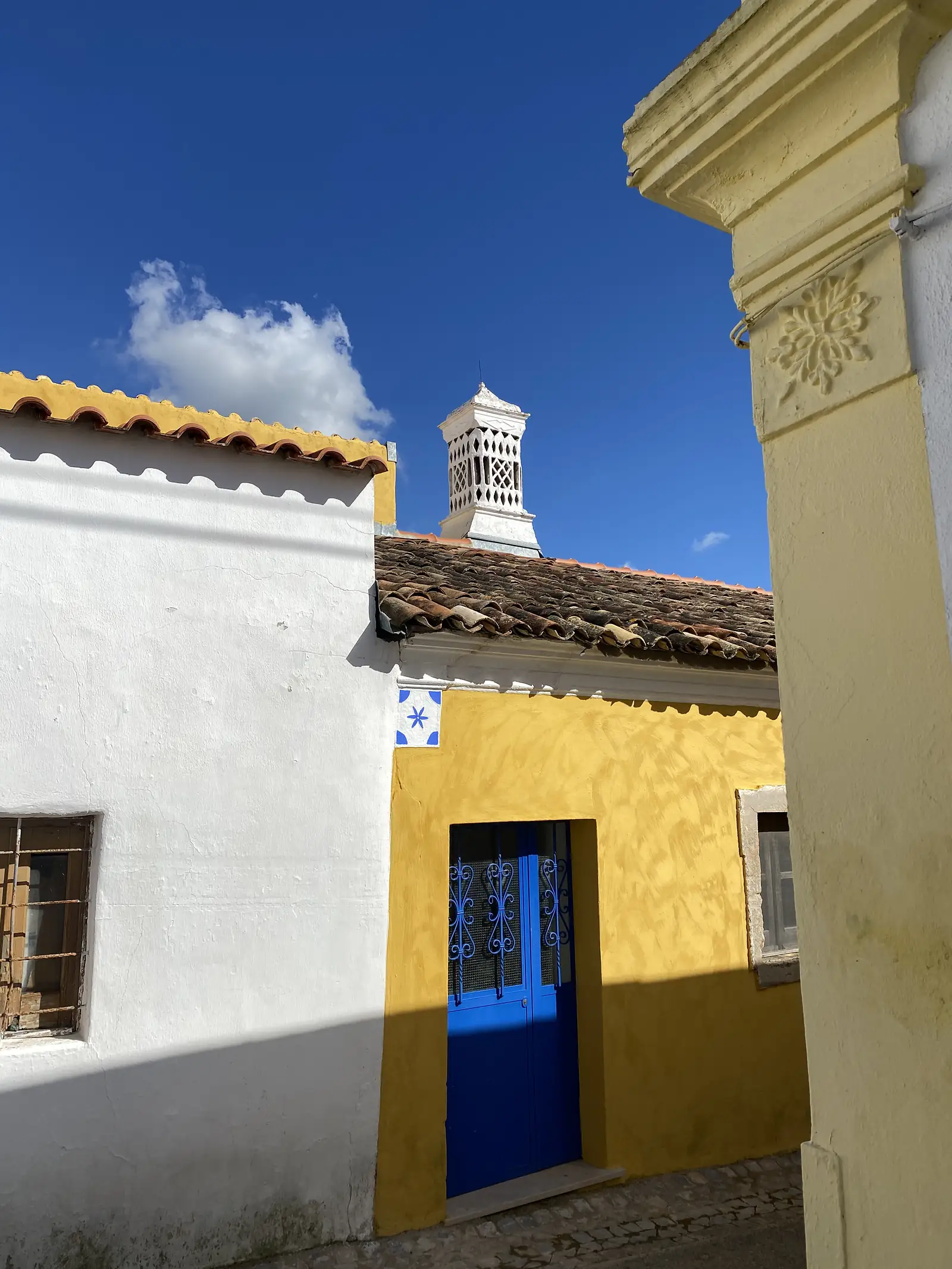 Corner of an ochre-washed cottage with a bright blue door behind wrought-iron grillework and a single blue-and-white tile above it, an ornate pierced white chimney standing against a deep blue sky.