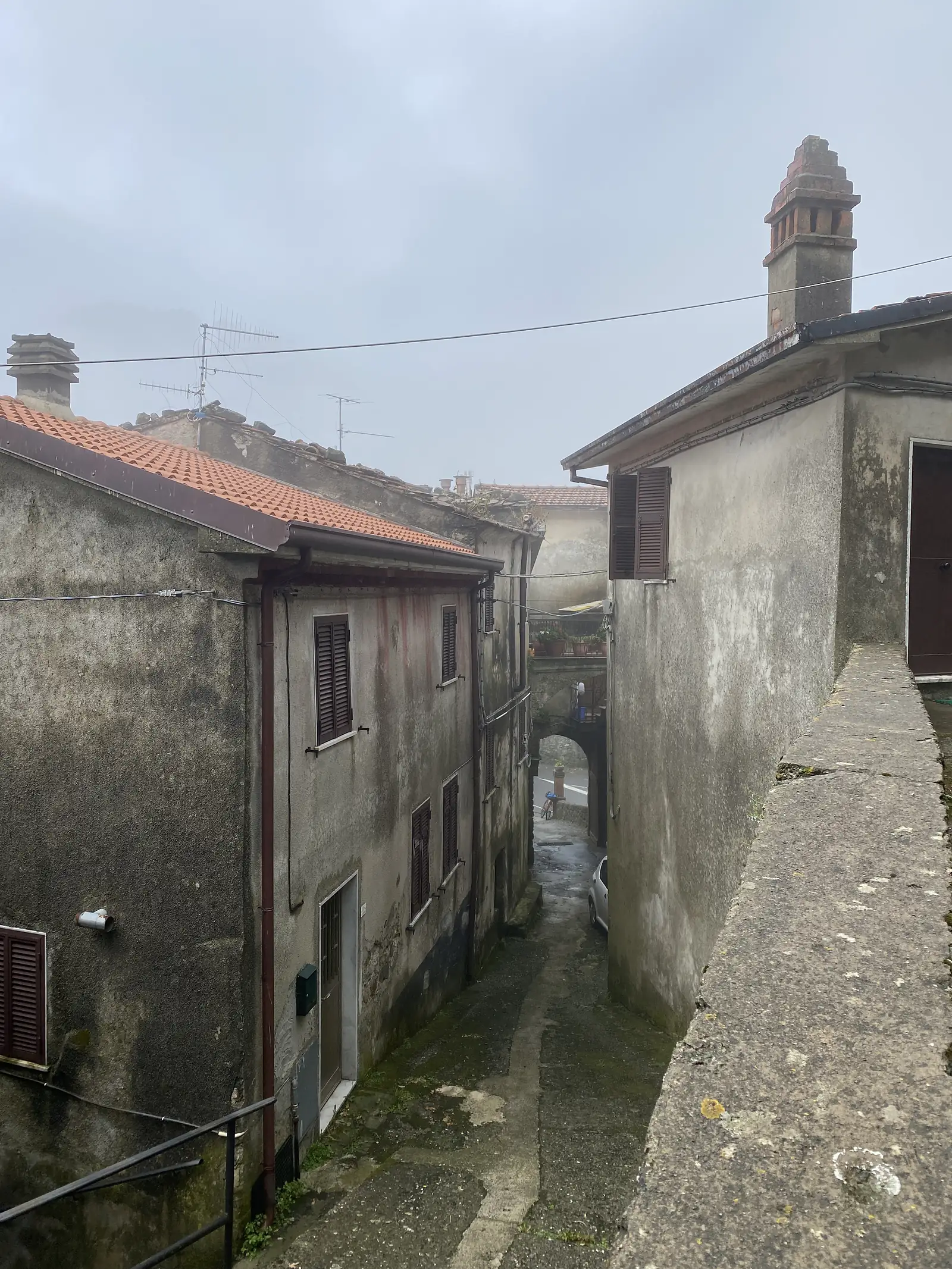 Terracotta-tiled rooftops, chimneys and roof vents crowd together above a narrow lane in a hill village, seen from a higher vantage point.