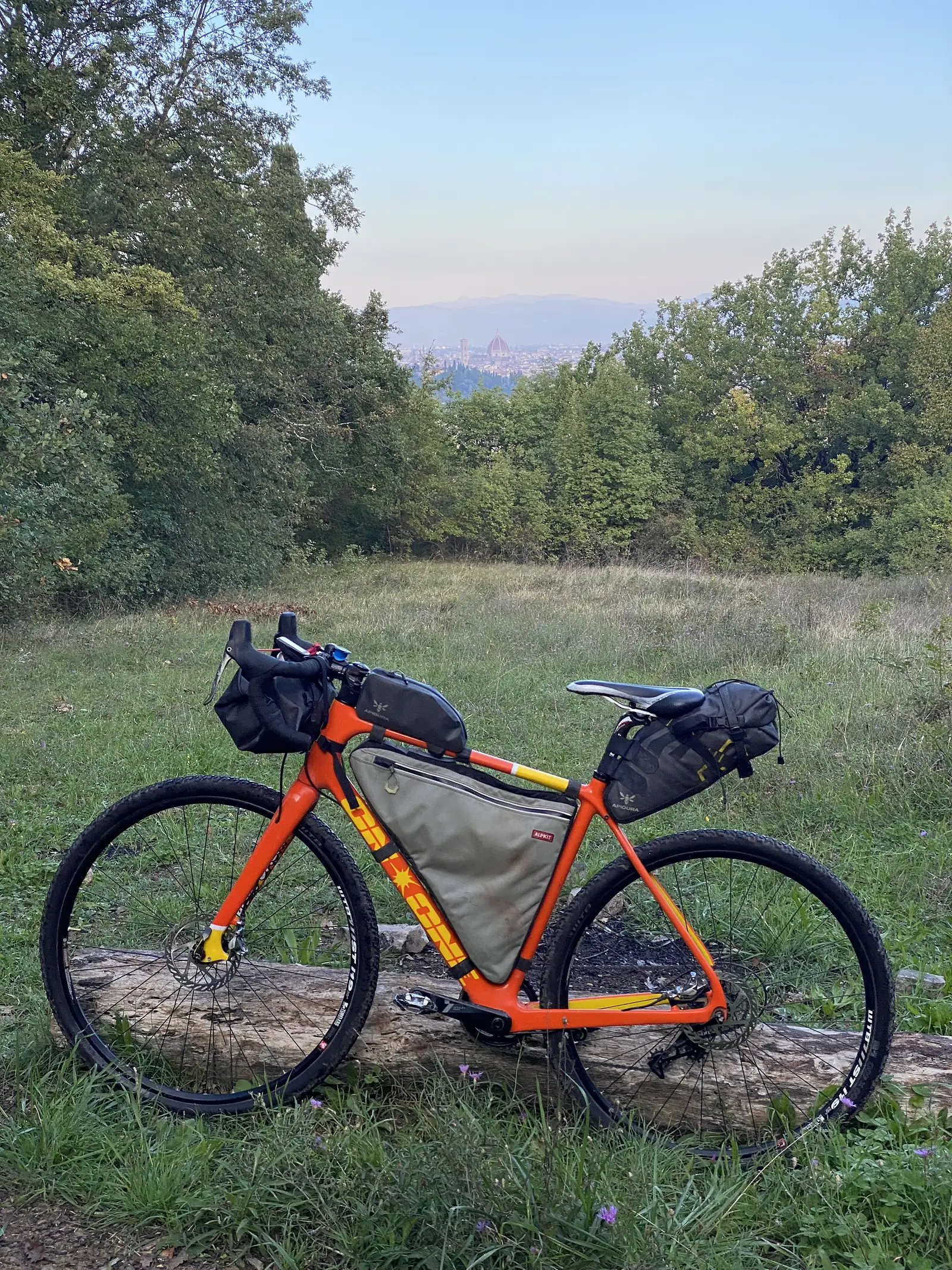A loaded orange bikepacking bike leaning against a wooden fence post on a grassy hillside track, with a hazy town visible in the valley below.