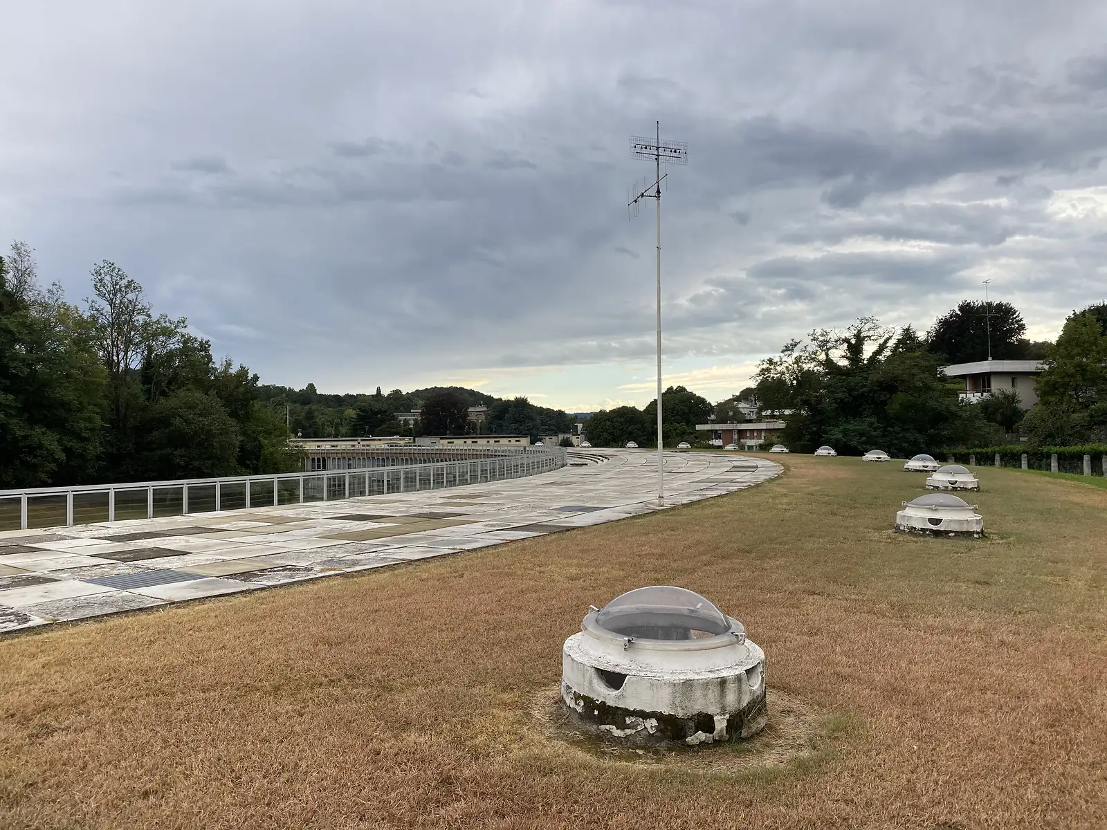 A domed rooflight set into dry grass on a rooftop terrace, with a tall aerial mast and a curved glazed building beyond, houses and hills visible in the distance.