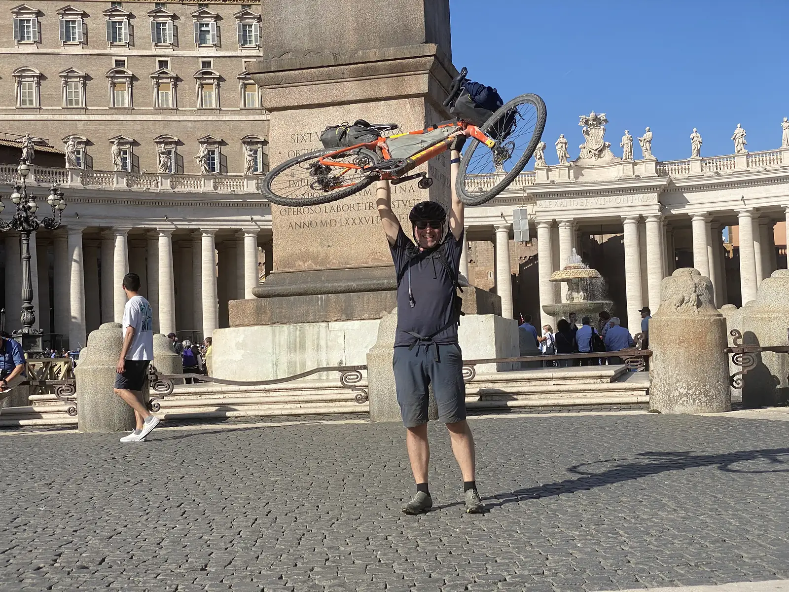 A cyclist hoists his loaded orange bike triumphantly overhead in St Peter's Square, the obelisk and Bernini's colonnade framing the scene behind him.