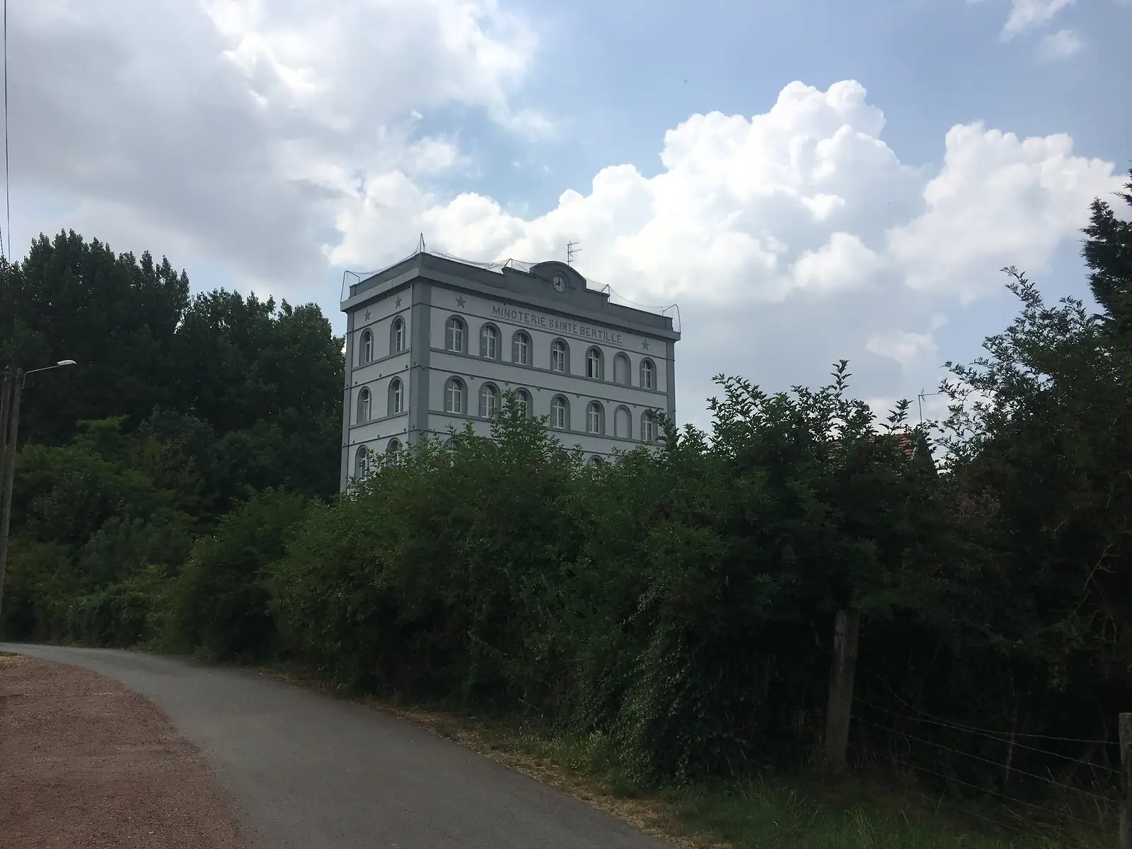 A tall grey industrial mill building labelled 'Minoterie Sainte Bertille' with a clock beneath its gable, rising above a hedgerow beside a country lane.