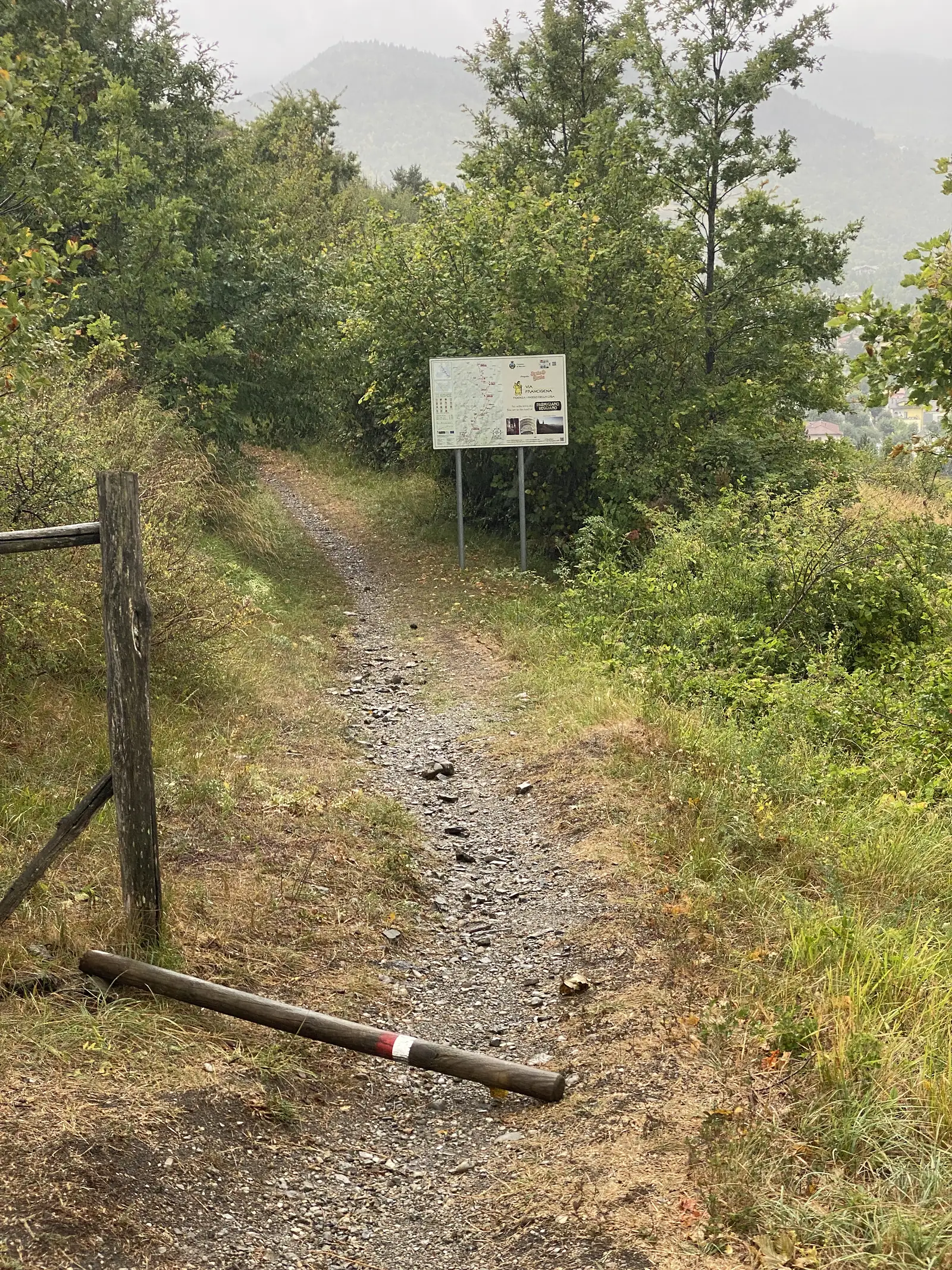 An information board illustrated with a route map and headed 'Via Francigena' stands beside a stony path bordered by a wooden rail and a red-and-white waymark post.