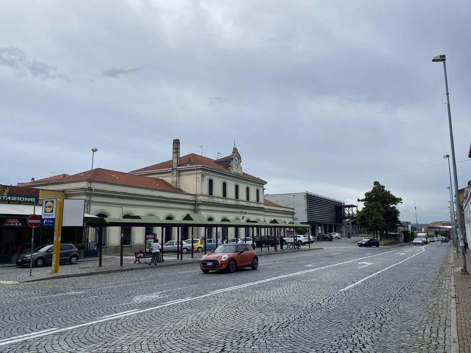 A former railway station building with a clock tower and terracotta roof, seen across a cobbled square past a red Mini car and a Lidl supermarket sign.