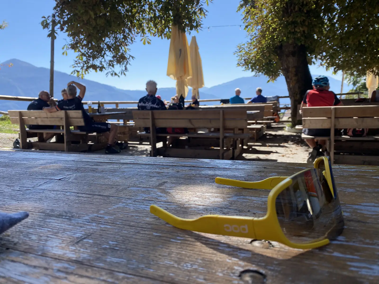 A pair of yellow wraparound cycling sunglasses lies on a weathered wooden table, with cyclists resting at benches under trees and mountains behind.