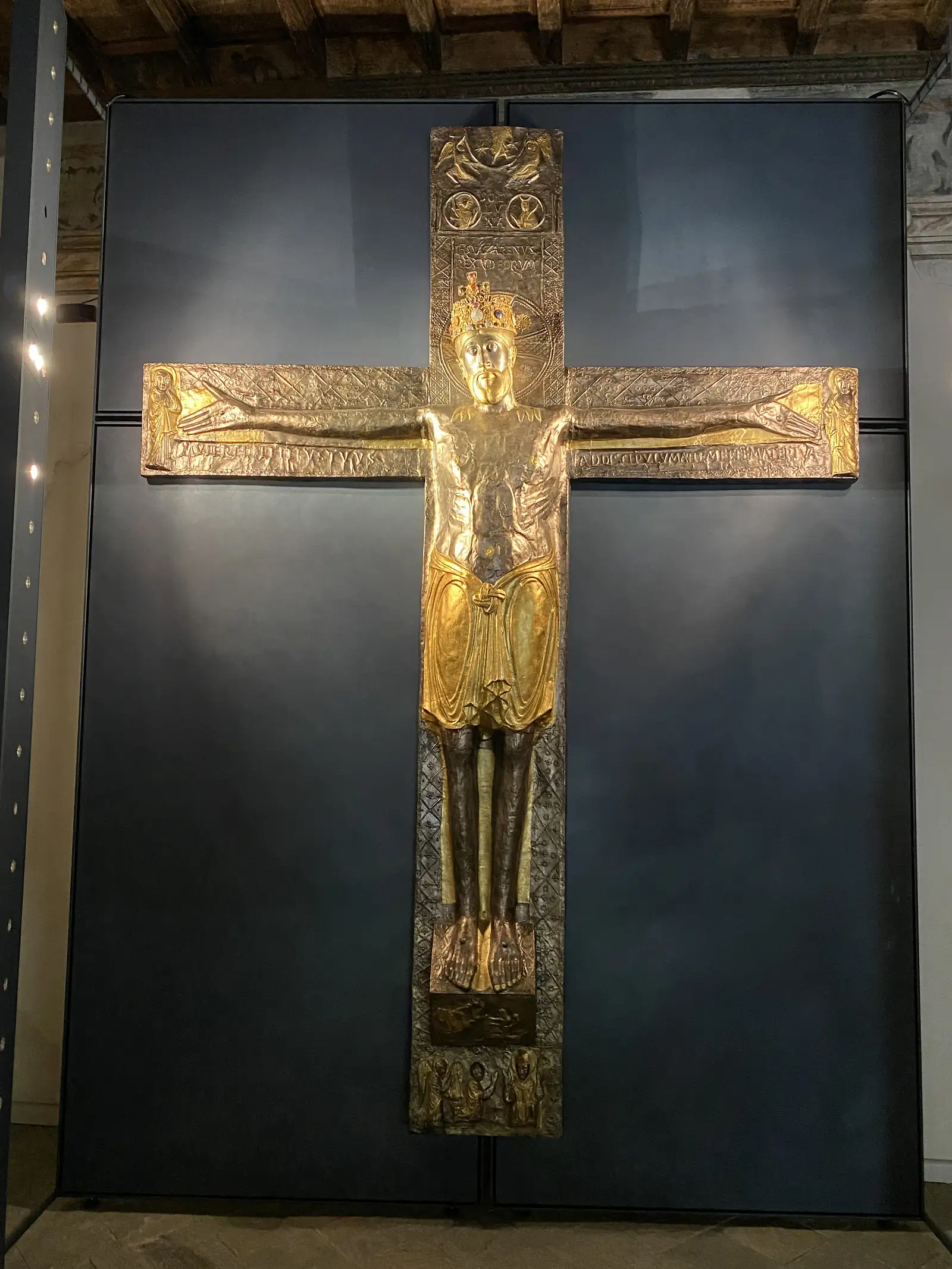 A gilded and silvered medieval reliquary cross bearing the raised figure of a crowned, robed Christ, displayed in a glass case against a dark blue backdrop.