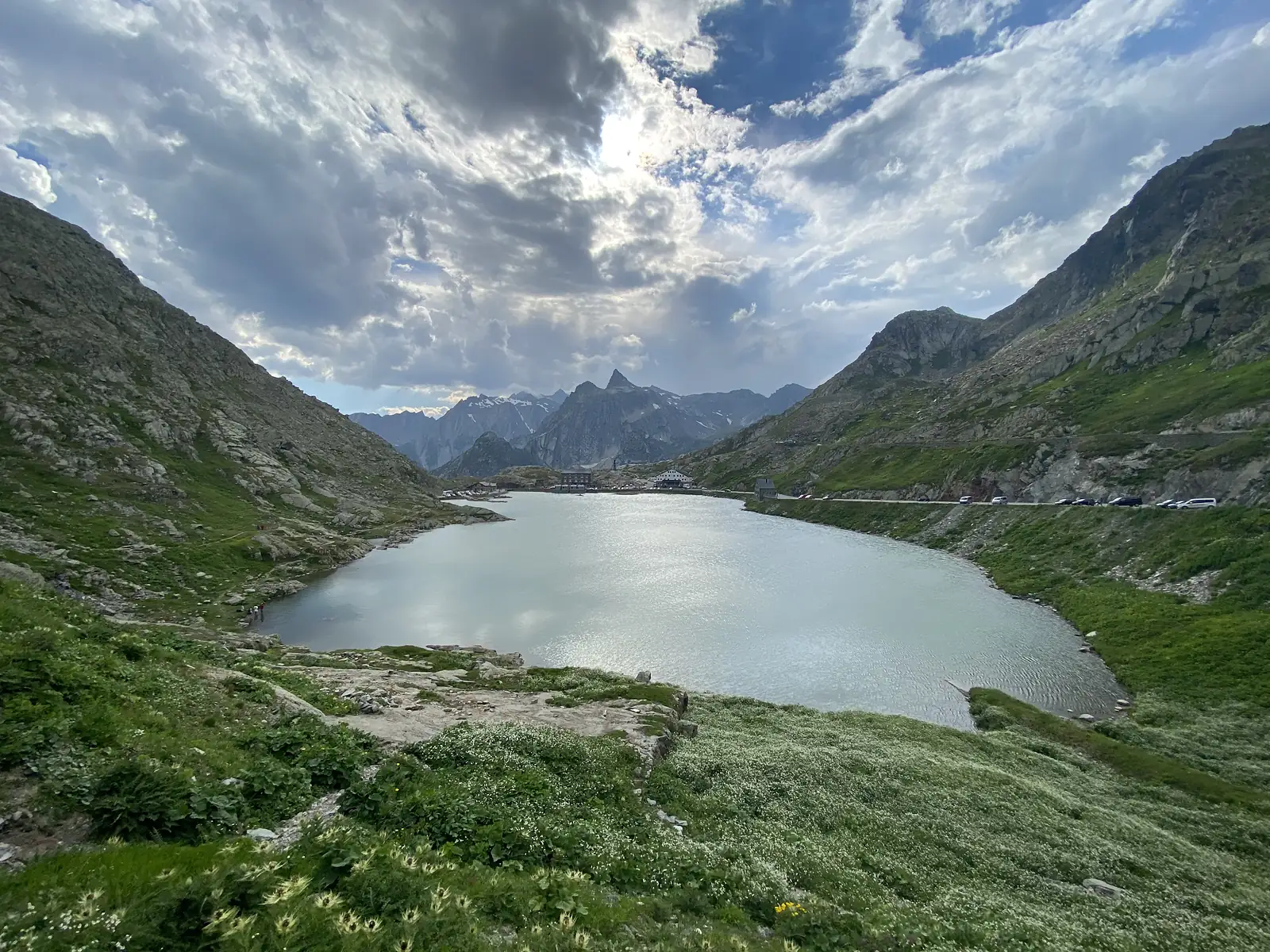 
A similar view of the pass lake under a dramatic, sun-pierced cloud bank, with a sharp peak rising beyond the hospice.
