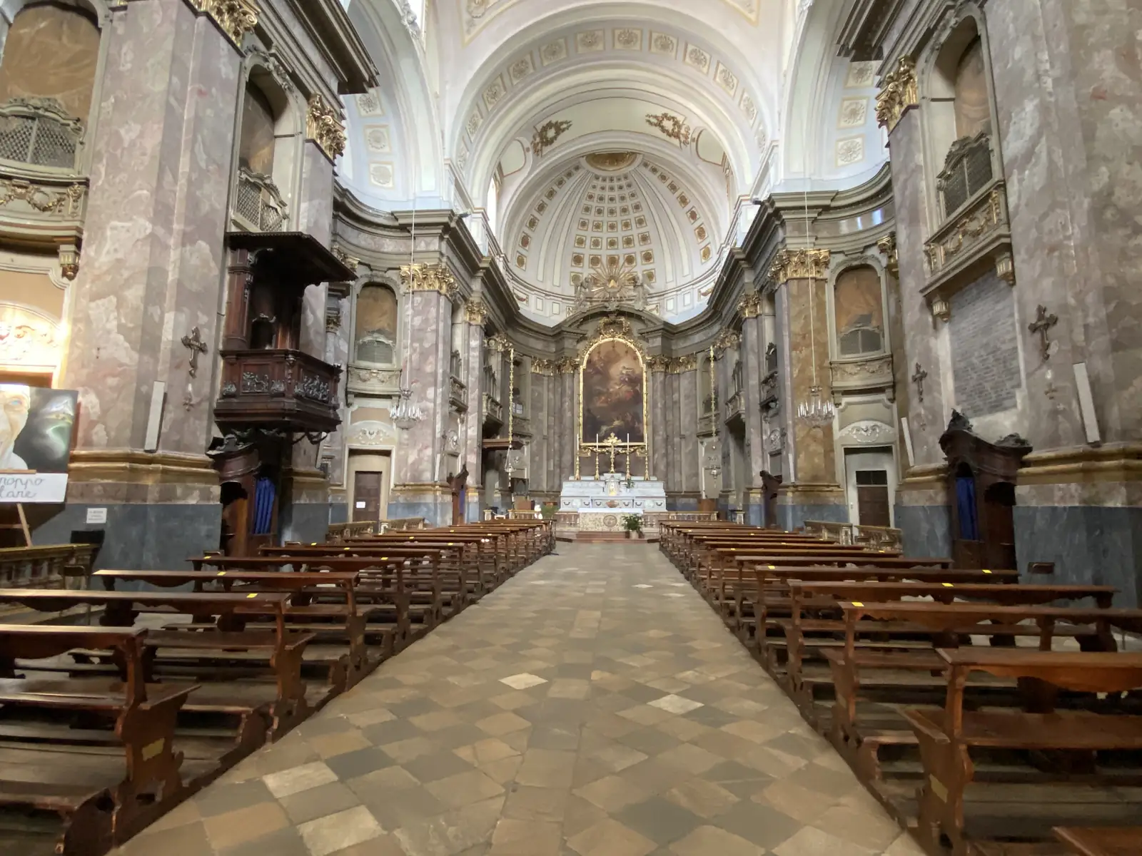 The barrel-vaulted nave of a Baroque church, its marble-clad pillars and painted dome leading the eye down empty wooden pews to a gilded altarpiece.