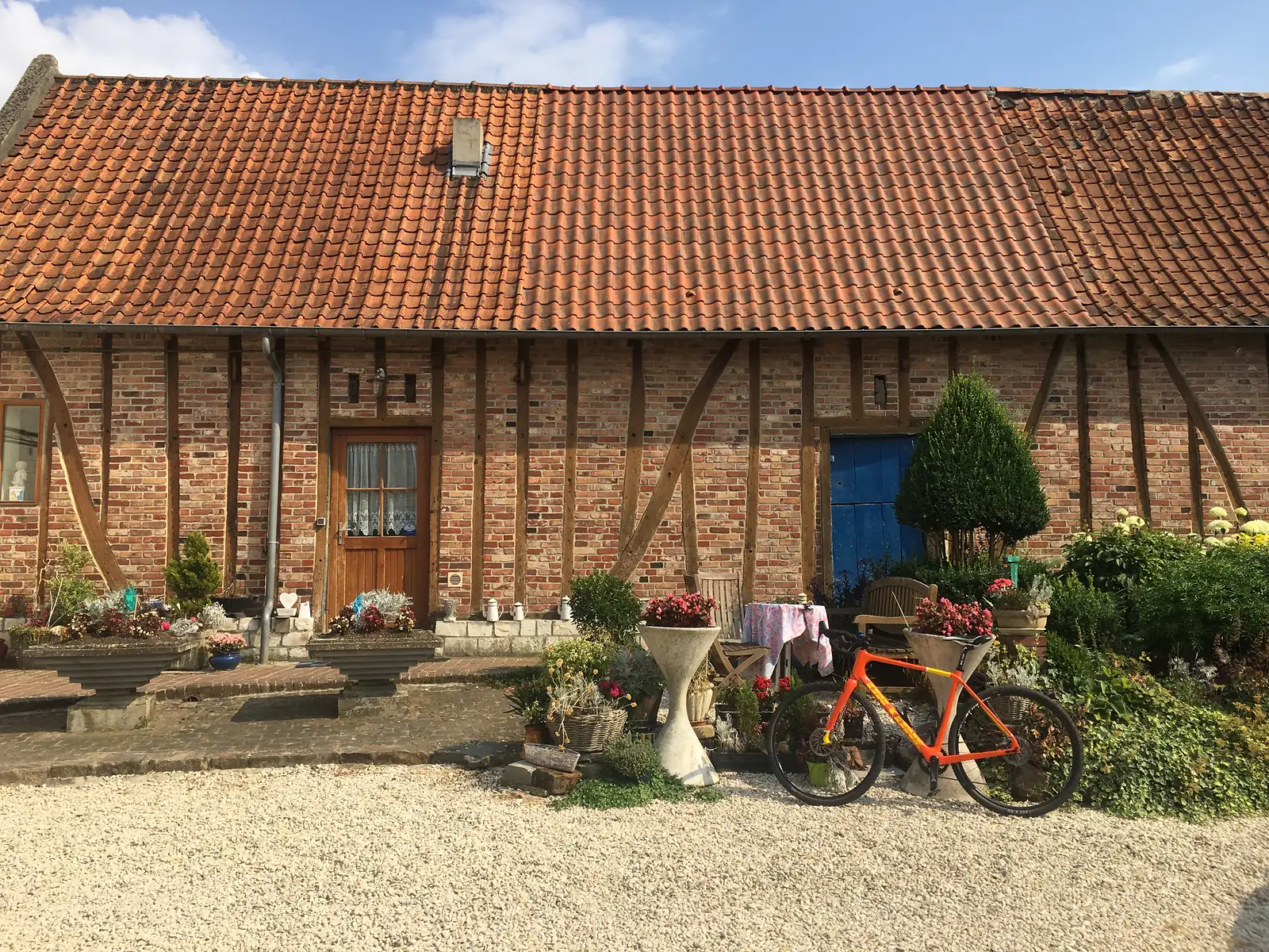 An orange gravel bike leans against a stone urn outside a half-timbered brick farmhouse, its doorway framed with pots of flowers.
