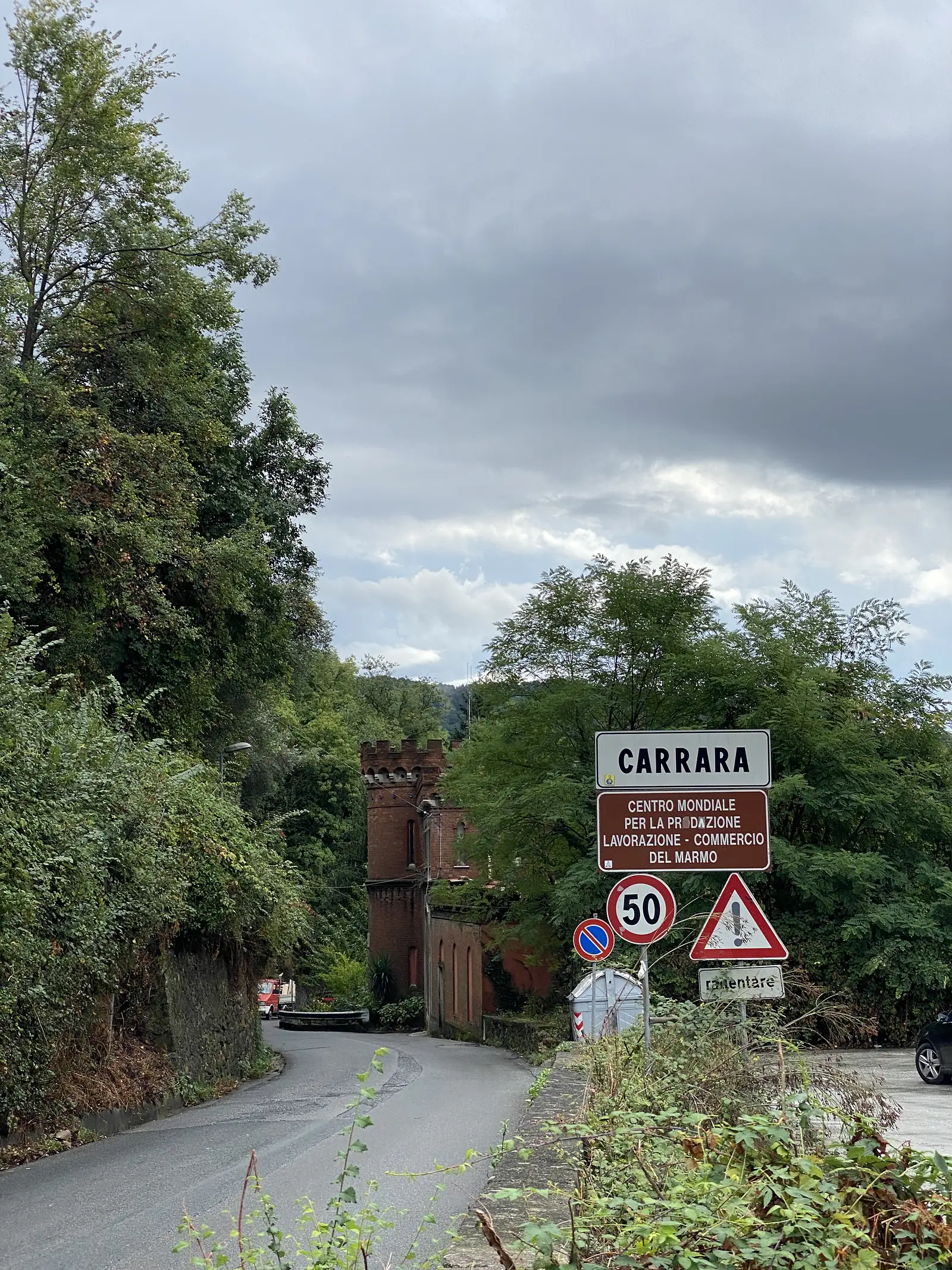 A road sign announcing Carrara as the world centre for the production, working and trade of marble, alongside a 50 km/h limit sign, with an old stone gateway visible through the trees behind.