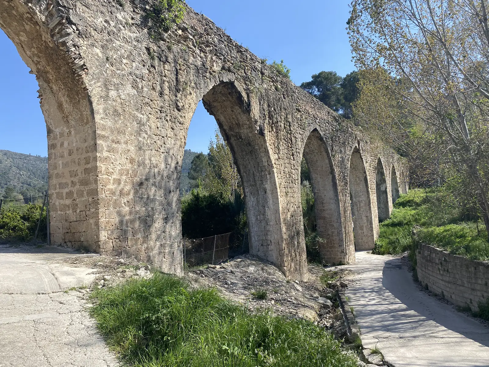 A tall stone aqueduct of pointed arches striding across a lane below a wooded hillside.