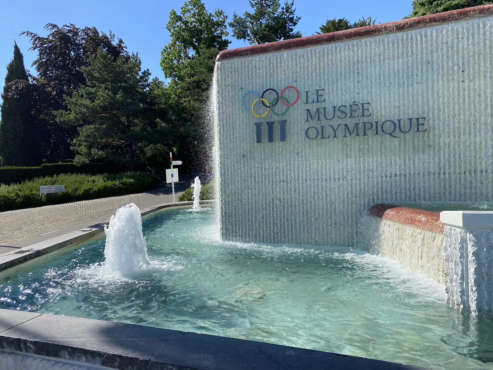 A stepped fountain cascading in front of the entrance wall of the Olympic Museum in Lausanne, its Olympic rings and lettering catching the sun.