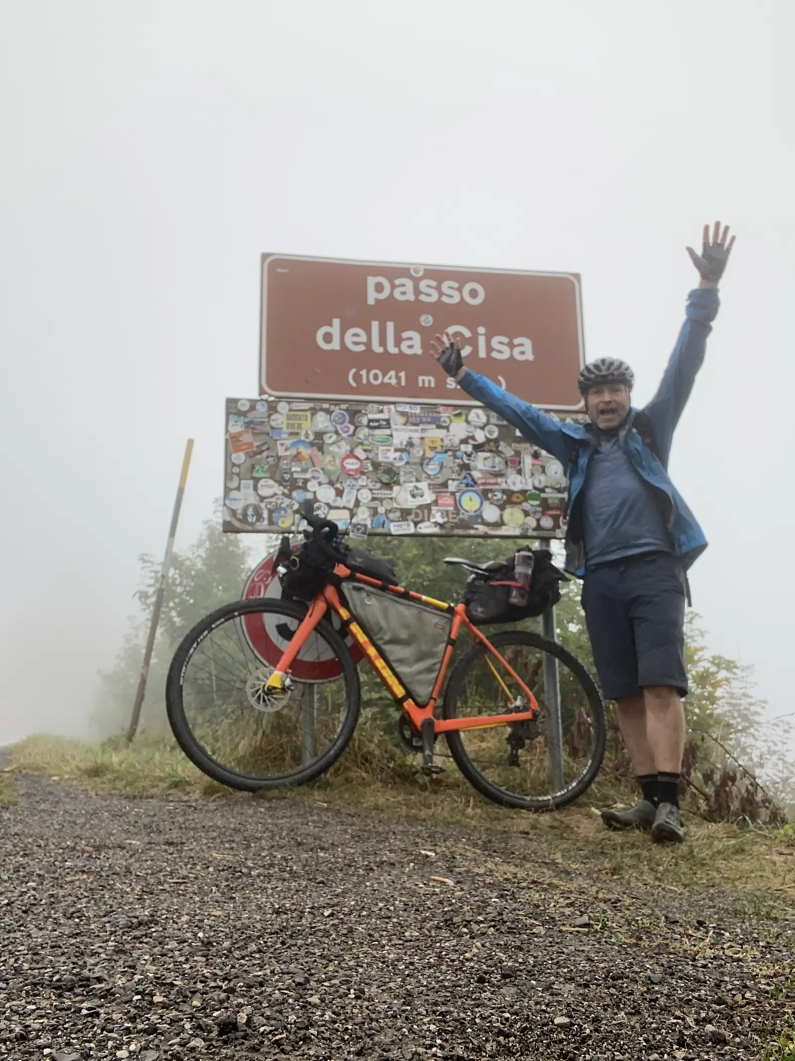 A cyclist in wet-weather gear throws his arms wide beside a laden orange bikepacking bike next to a brown sign reading Passo della Cisa (1041 m), fog blotting out the view behind.