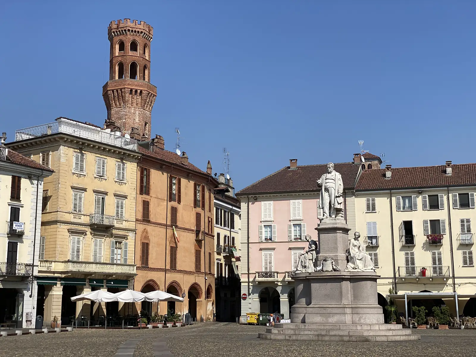 A crenellated brick tower rises above a piazza in Vercelli, where a statue of Camillo Cavour stands on a pedestal before pastel-coloured townhouses.