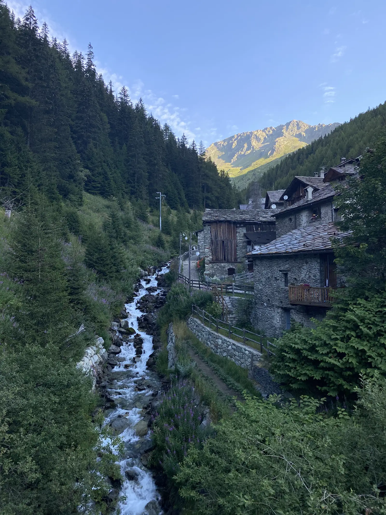 A mountain stream tumbles over rocks past a row of stone-built houses with slate roofs, a sunlit peak visible at the head of the valley beyond.